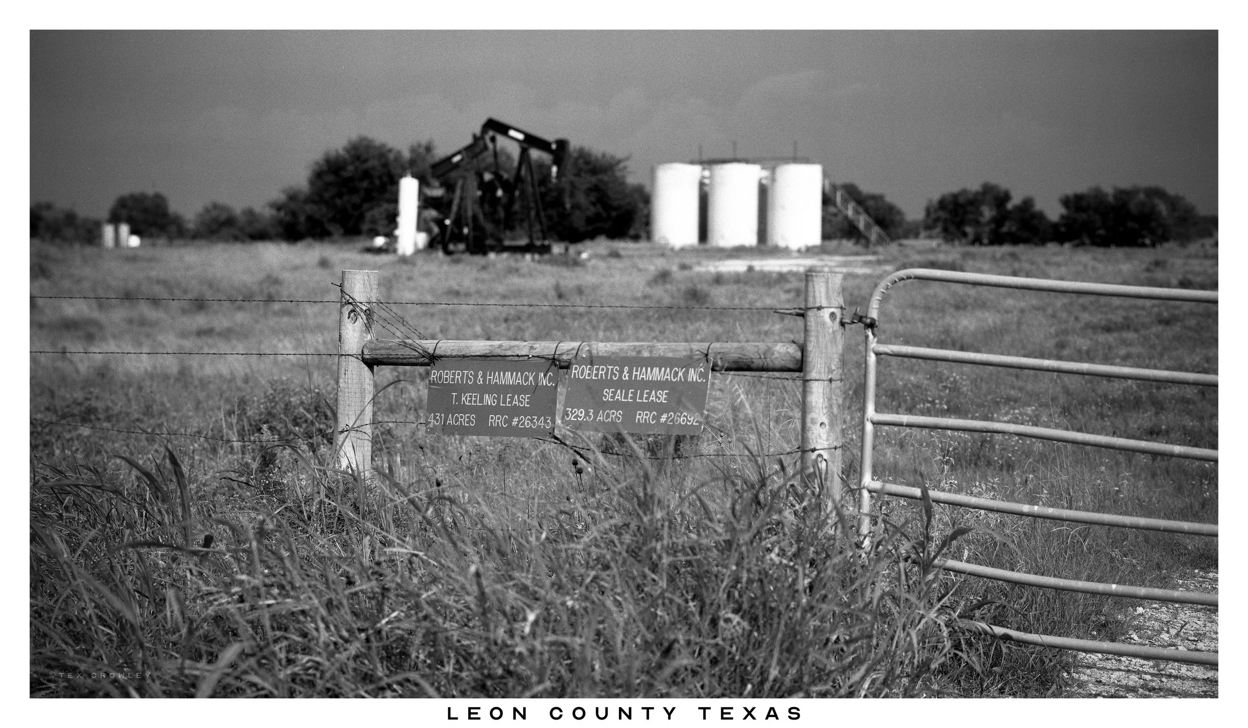 Fenced farmland with signs indicating lease agreements, with oil pumpjack and storage tanks in the background, in Leon County, Texas.