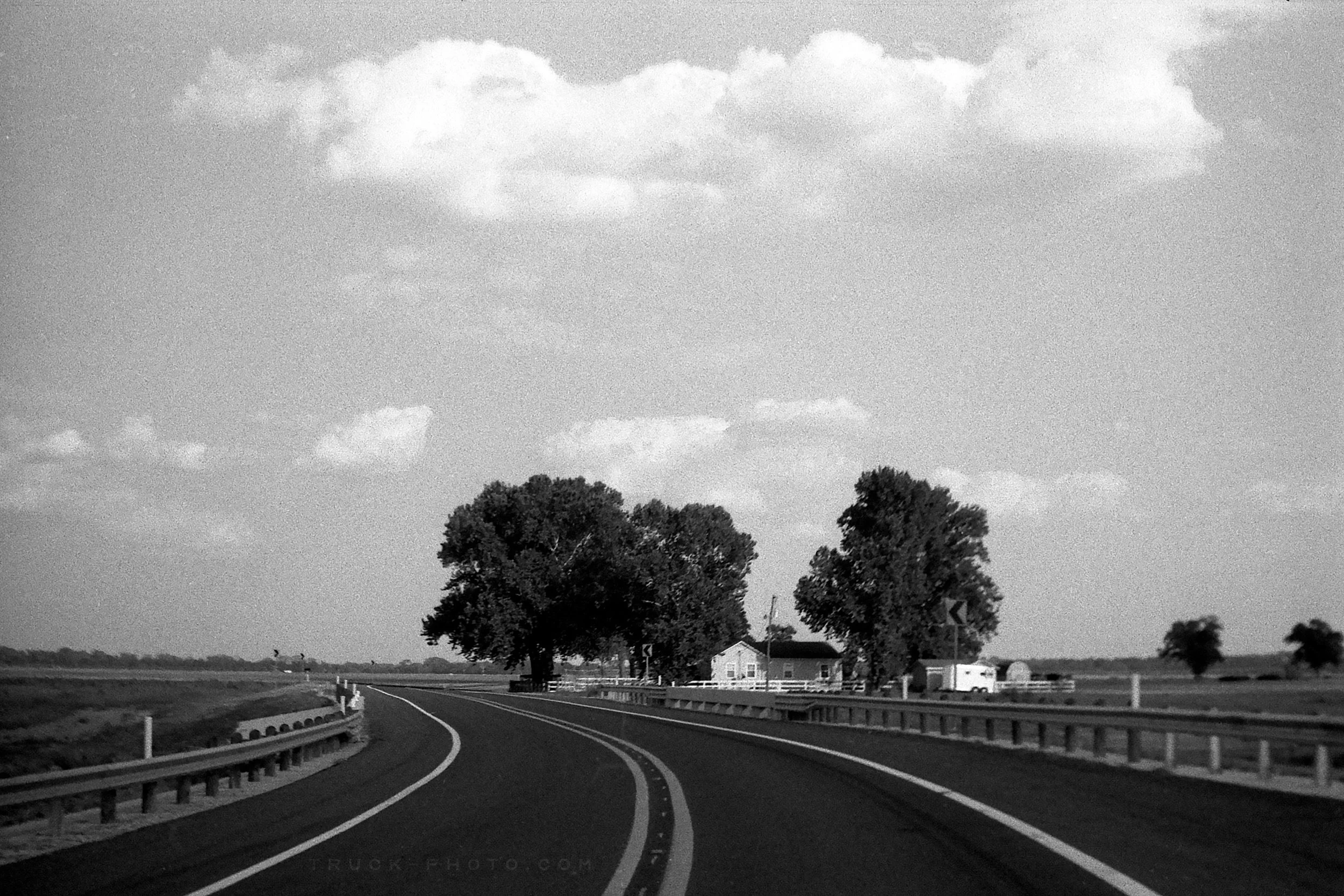 Black and white photo of a rural highway curve with guardrails, trees, a house, and a trailer in the background under a partly cloudy sky.