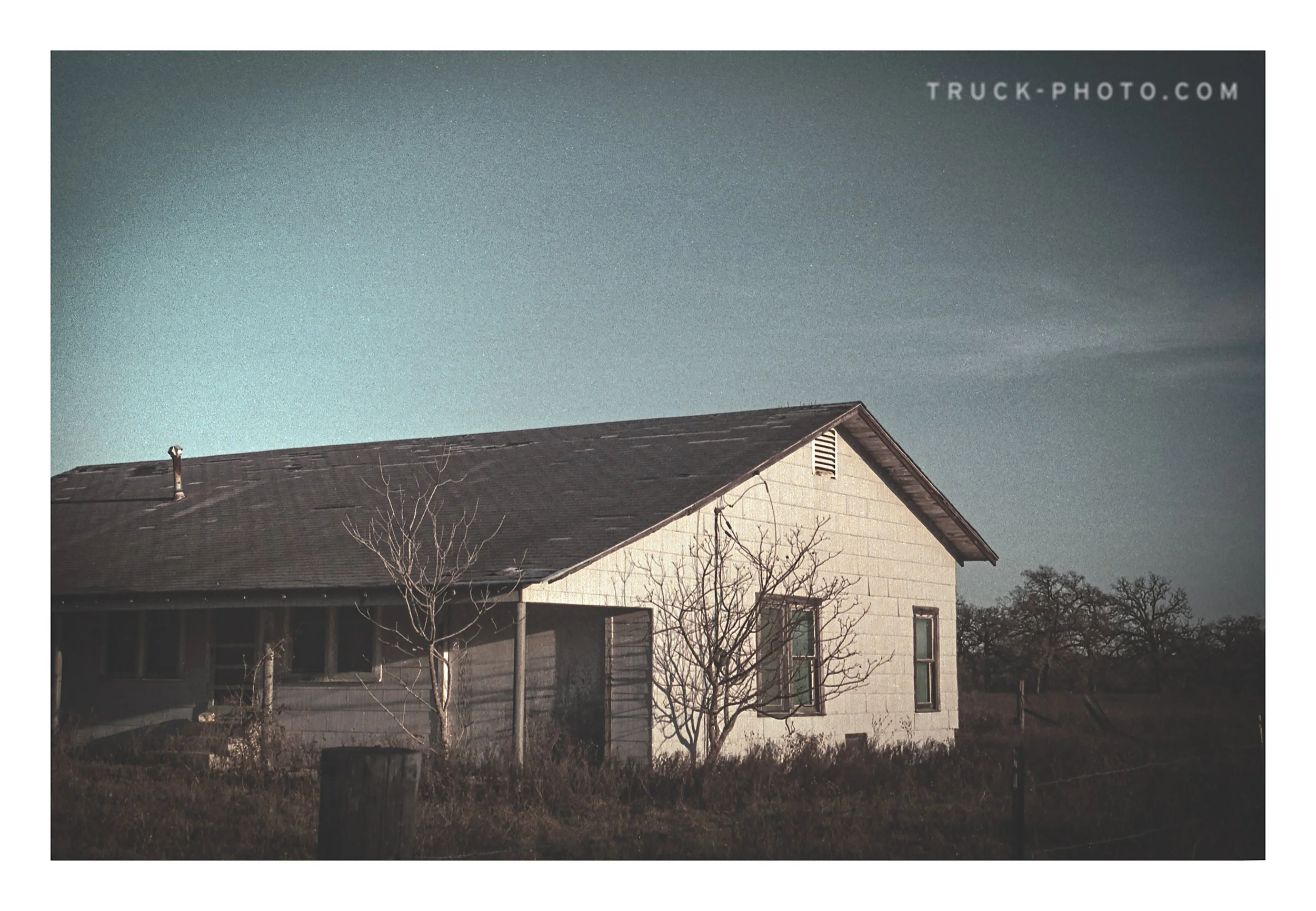 A small house with a gray roof and white exterior walls, surrounded by sparse trees and dry grass, under a clear sky.