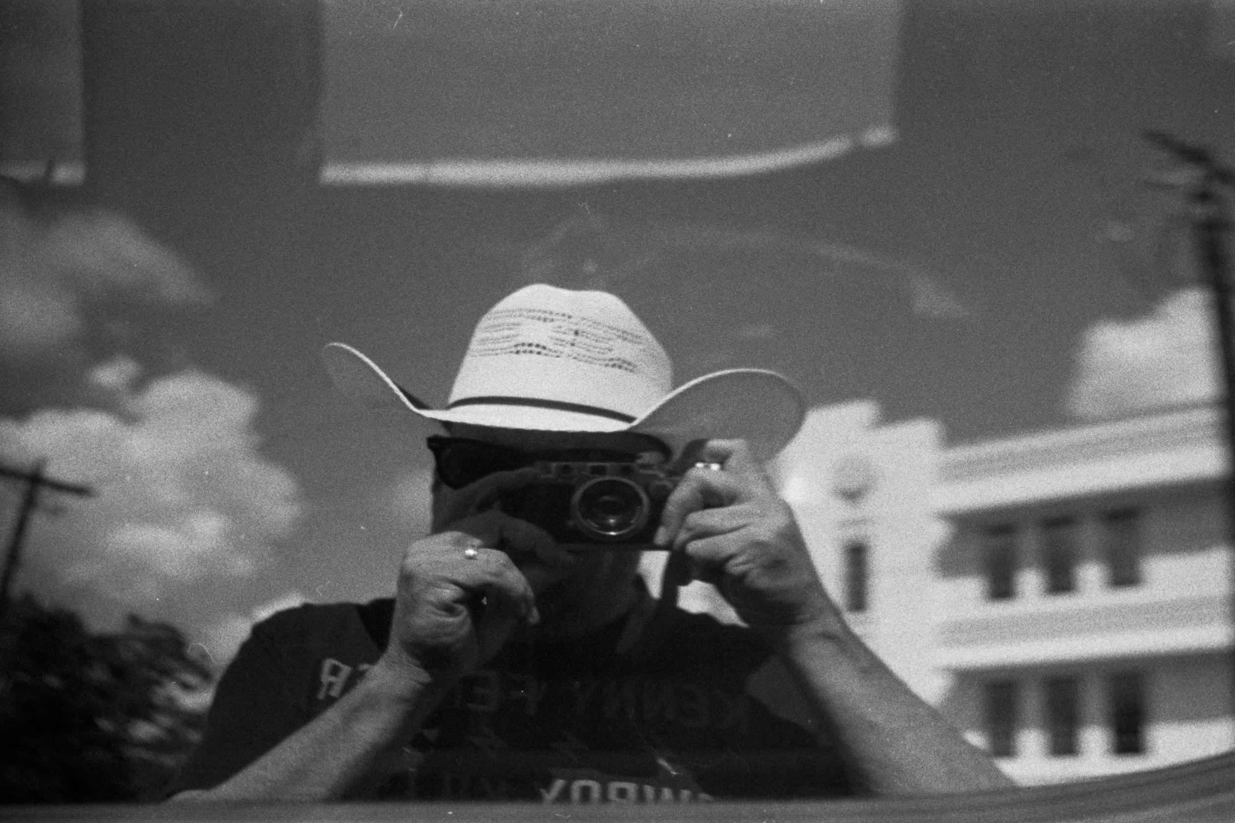 A person taking a photograph of their reflection in a shiny surface, wearing a cowboy hat and holding a camera with both hands.