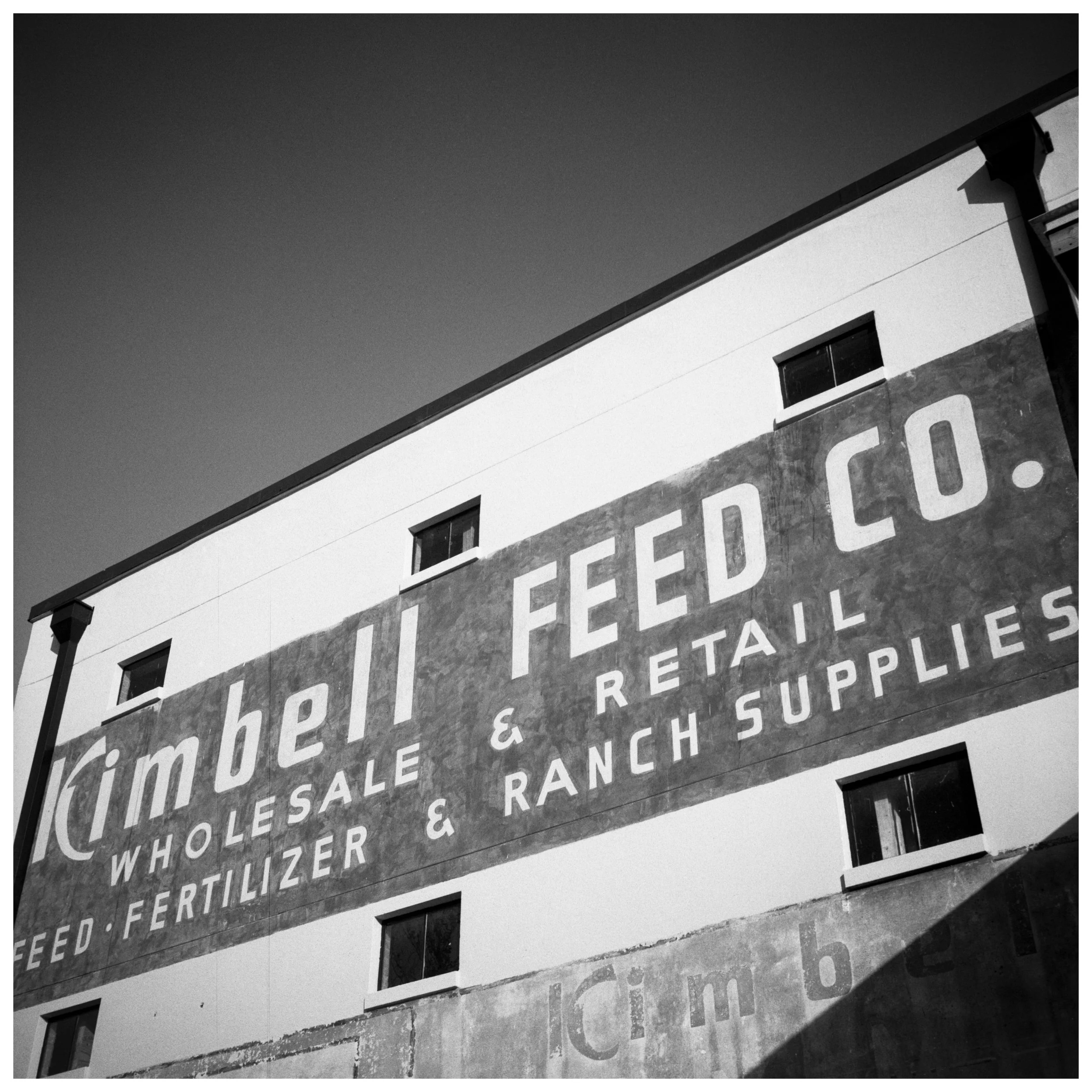 A large building with a sign that reads 'Kimbert Feed Co. Wholesale & Retail - Feed, Fertilizer, Ranch Supplies' in white letters on a weathered dark background.