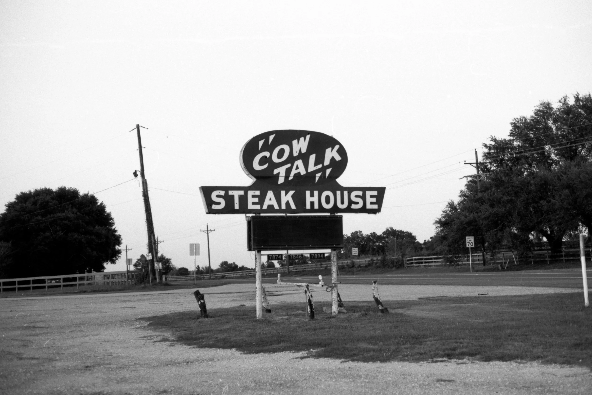 Black and white photo of a roadside sign for Cow Talk Steak House with a blinking electronic message board underneath, surrounded by trees and power lines.
