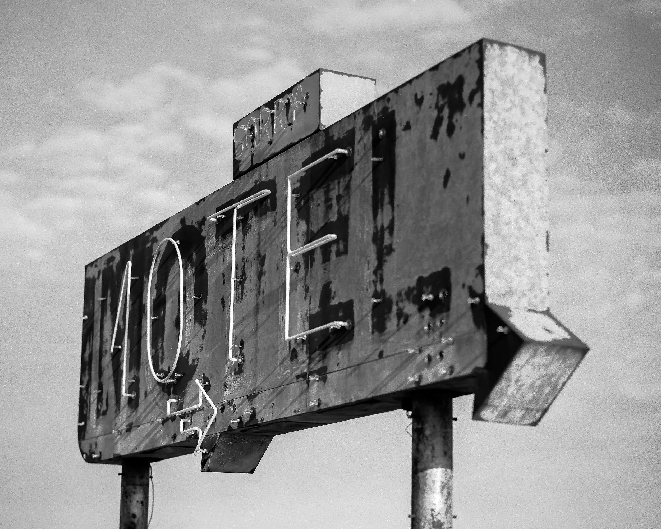 A weathered, vintage sign that reads ' HOTEL' with neon lights, mounted on metal poles against a cloudy sky.