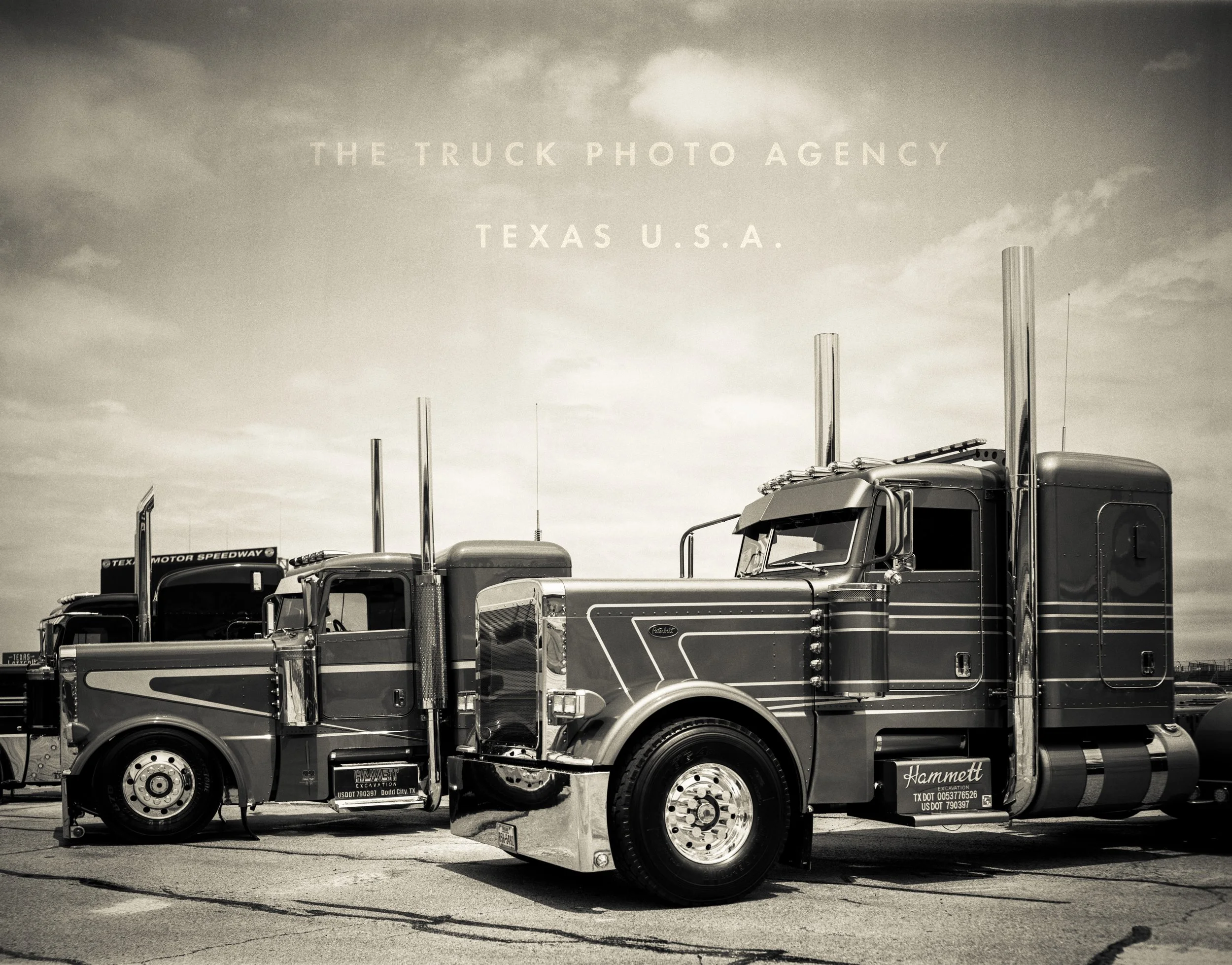 Black and white photo of two large semi-trucks parked side by side at Texas Motor Speedway in Texas, U.S., with a cloudy sky overhead.