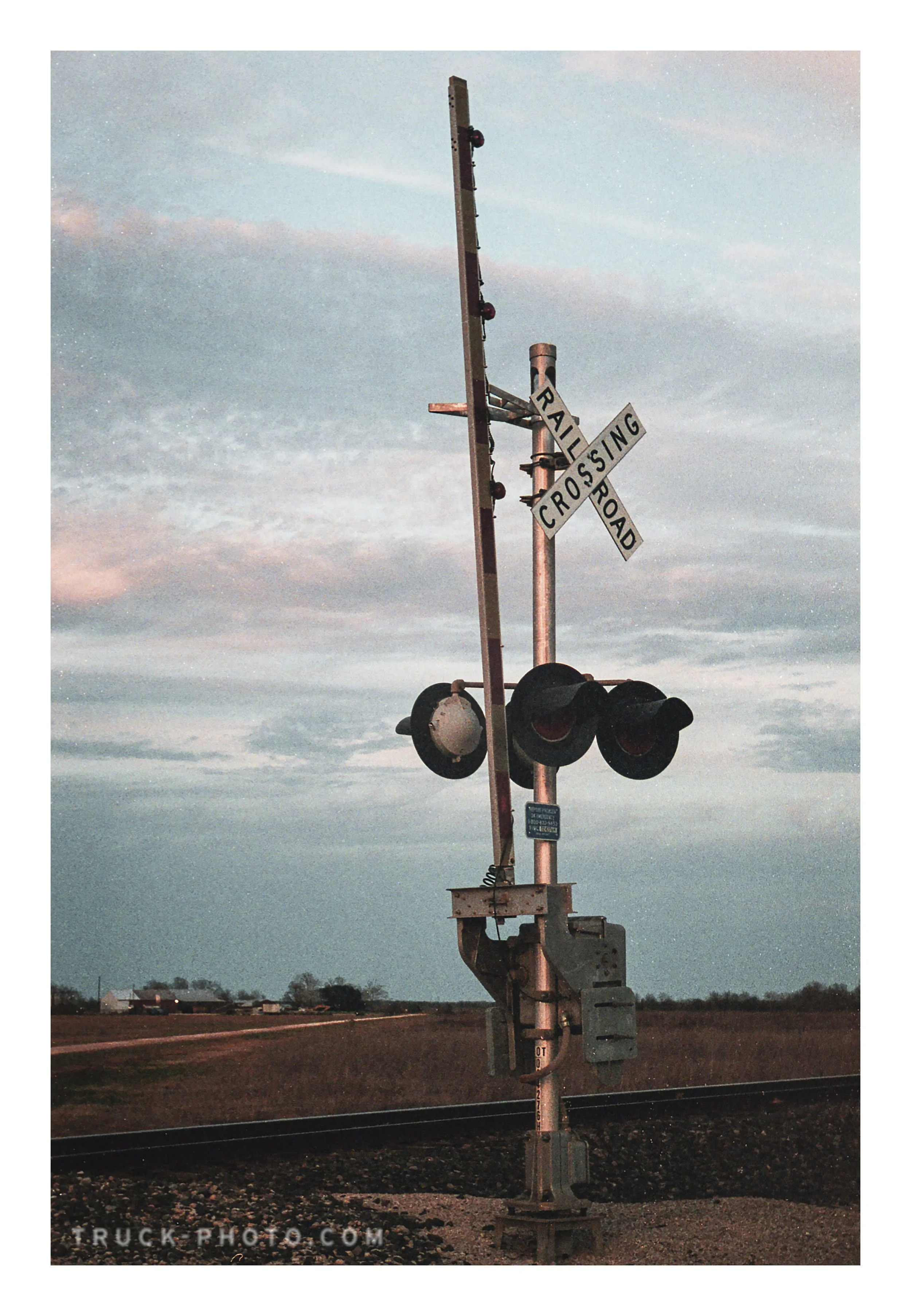 A train crossing signal with two circular red lights, a crossing sign, and a barrier arm near railroad tracks in a rural area during sunset or dawn.