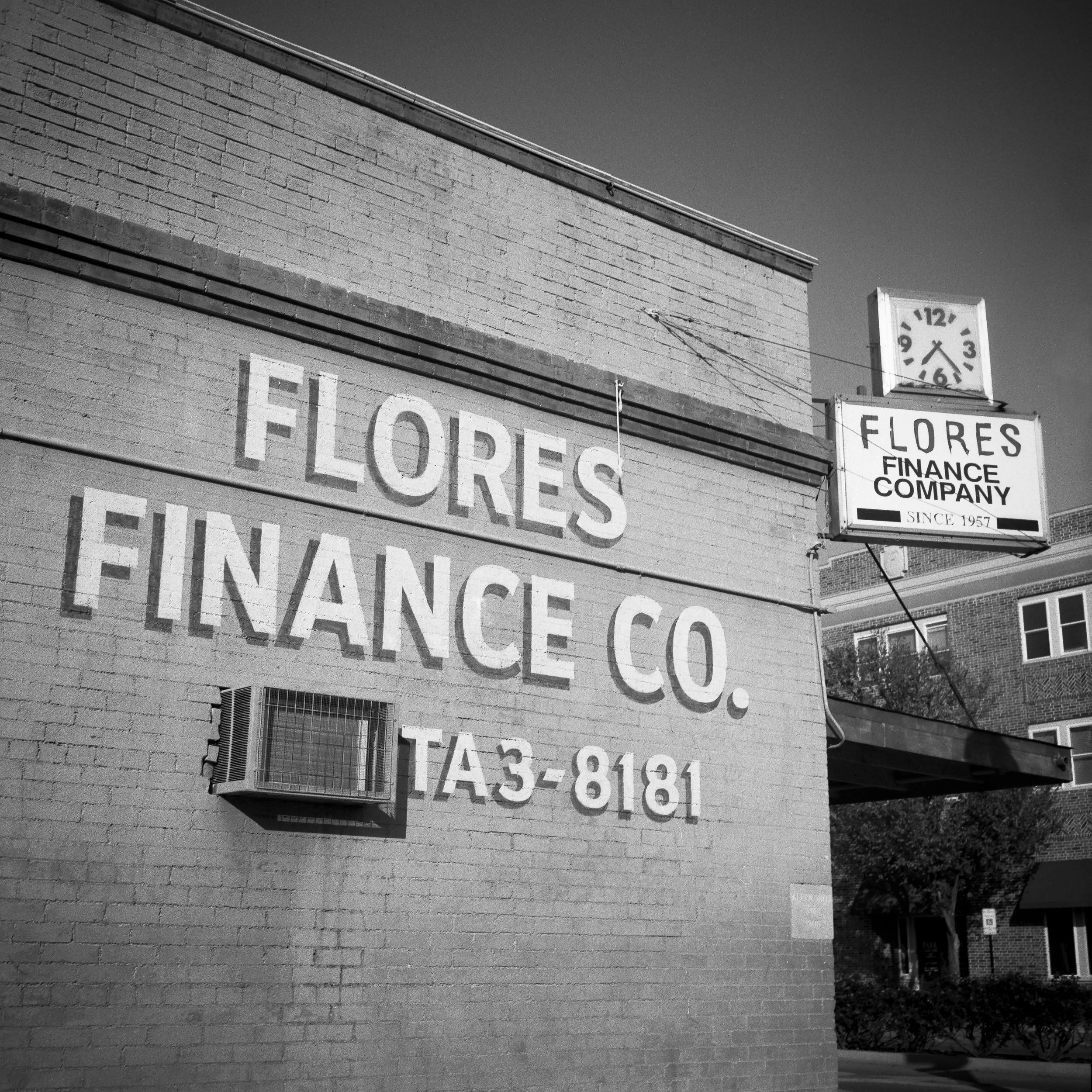 Black and white photo of a brick building with large painted sign reading "FLORES FINANCE CO." and a phone number "TA 3-8181". There is a clock on top of a sign showing around 2:07, with the same business name and a note "SINCE 1957".
