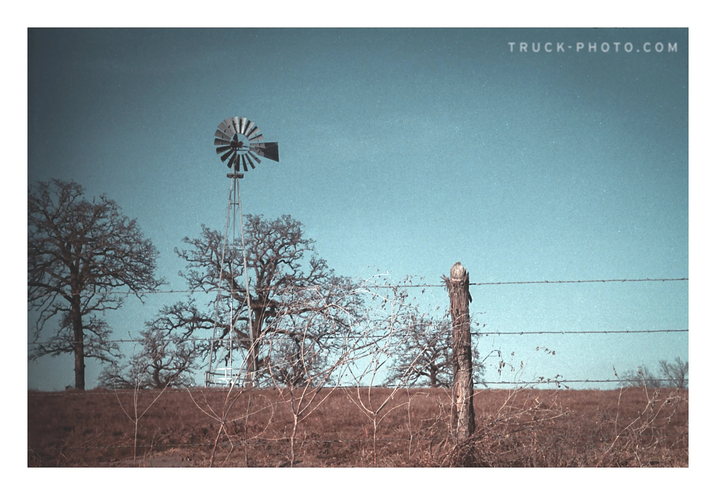 A rural scene featuring a windmill, barbed wire fence, and leafless trees under a clear blue sky.
