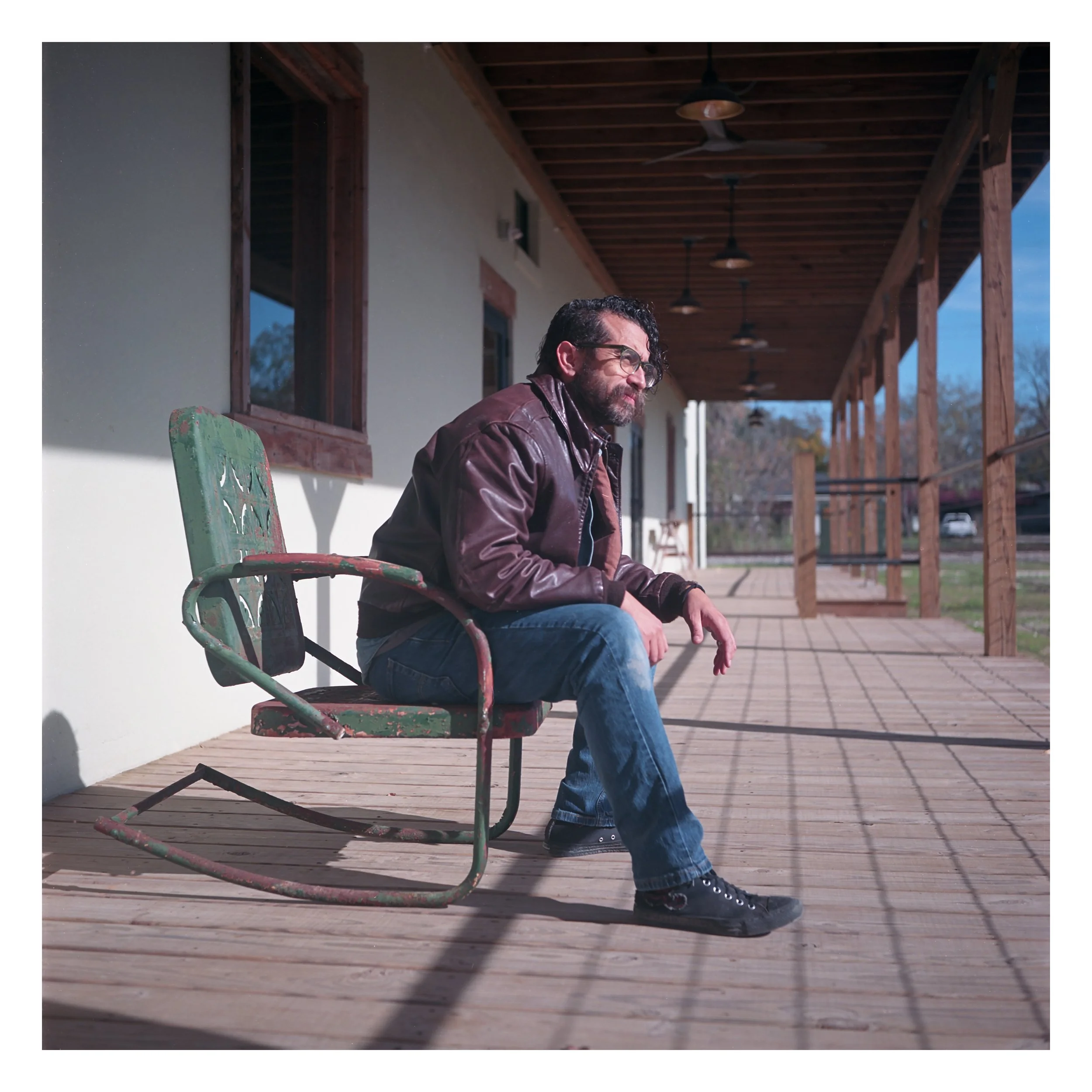 A man with glasses, beard, and curly hair sitting on a weathered green rocking chair on a wooden porch. He is wearing a brown leather jacket, jeans, and black sneakers, and appears to be looking down or lost in thought.