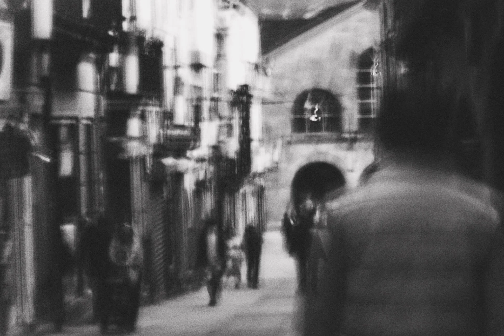 Black and white photo of an urban street scene with apartment buildings, people walking on the sidewalk, and an arched doorway in the wall.