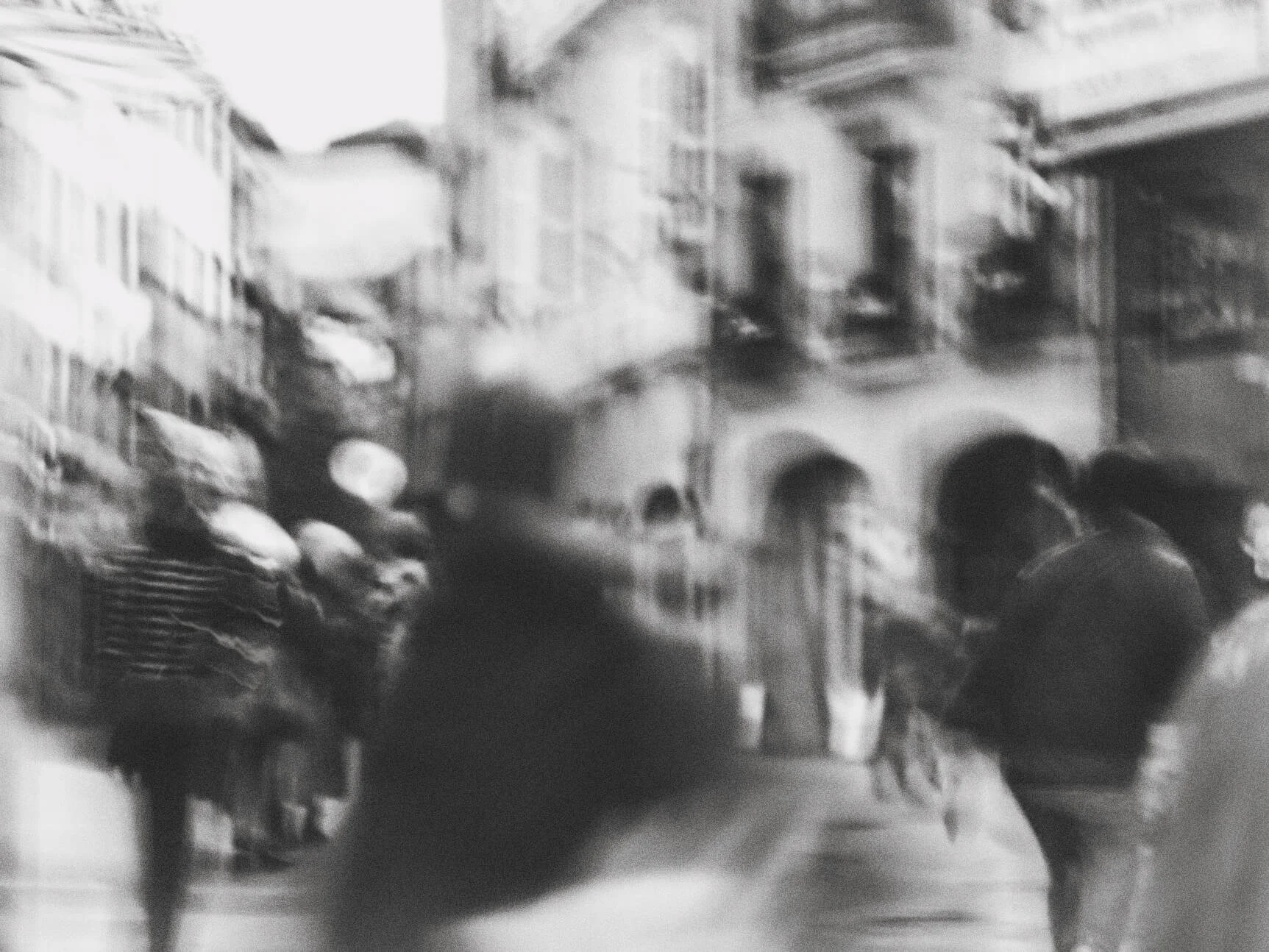 Blurred black and white photo of a city street with several people walking and buildings in the background.