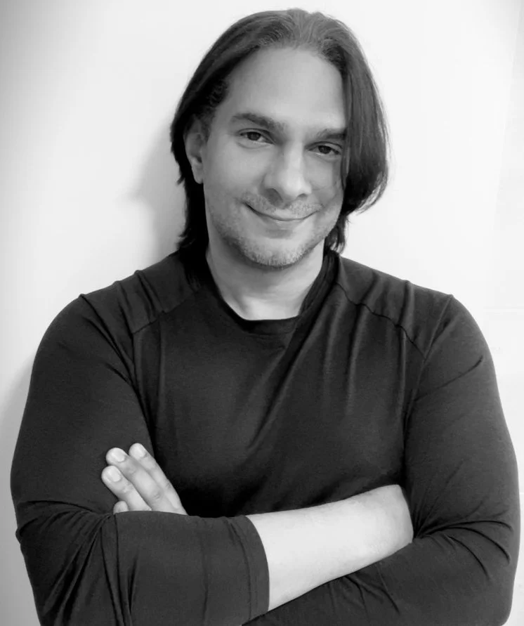 Black-and-white portrait of a man with long hair, smiling, in a black long-sleeve shirt, with arms crossed