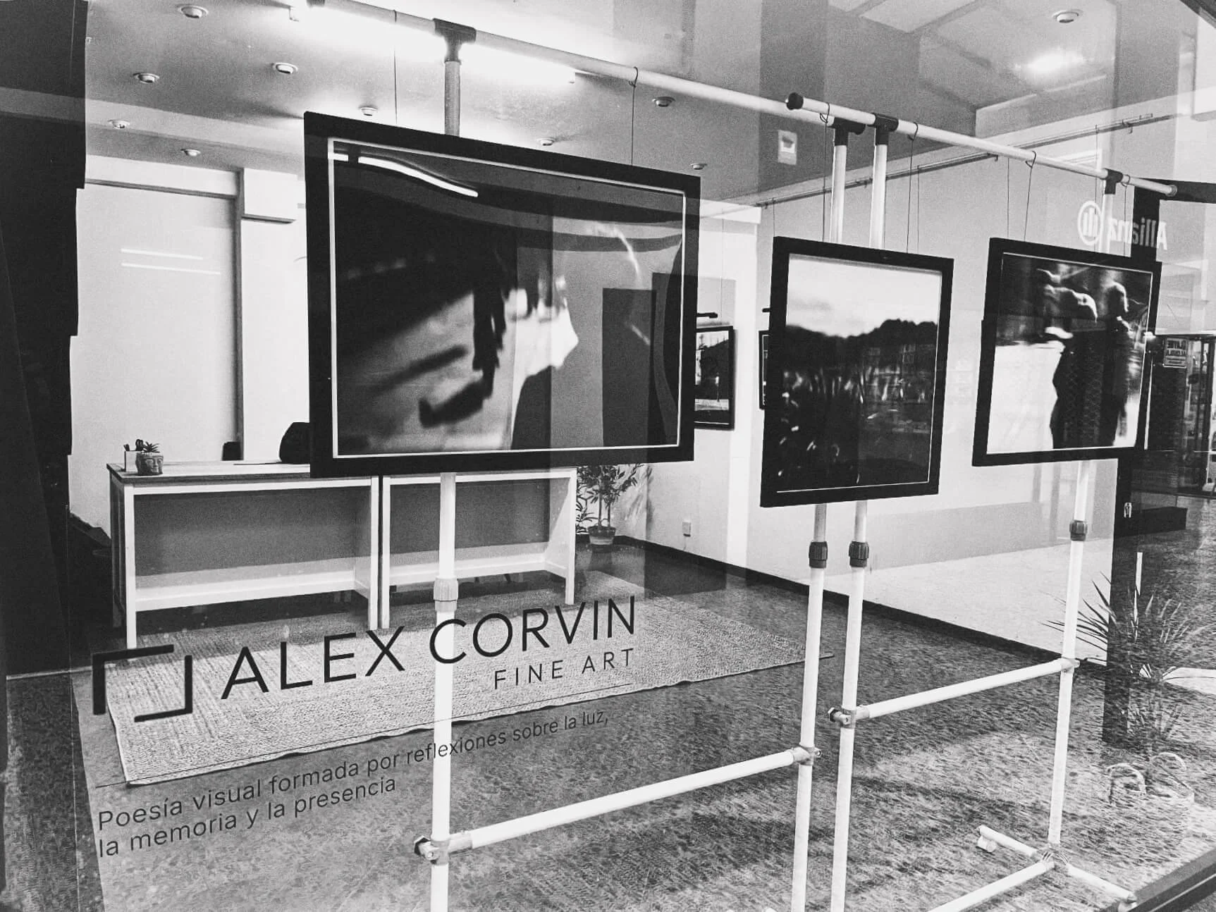 Black and white photograph of an art gallery display window featuring framed photographs. The gallery sign reads "ALEX CORVIN FINE ART" and has a description in Spanish about visual poetry and reflection on light, memory, and presence.