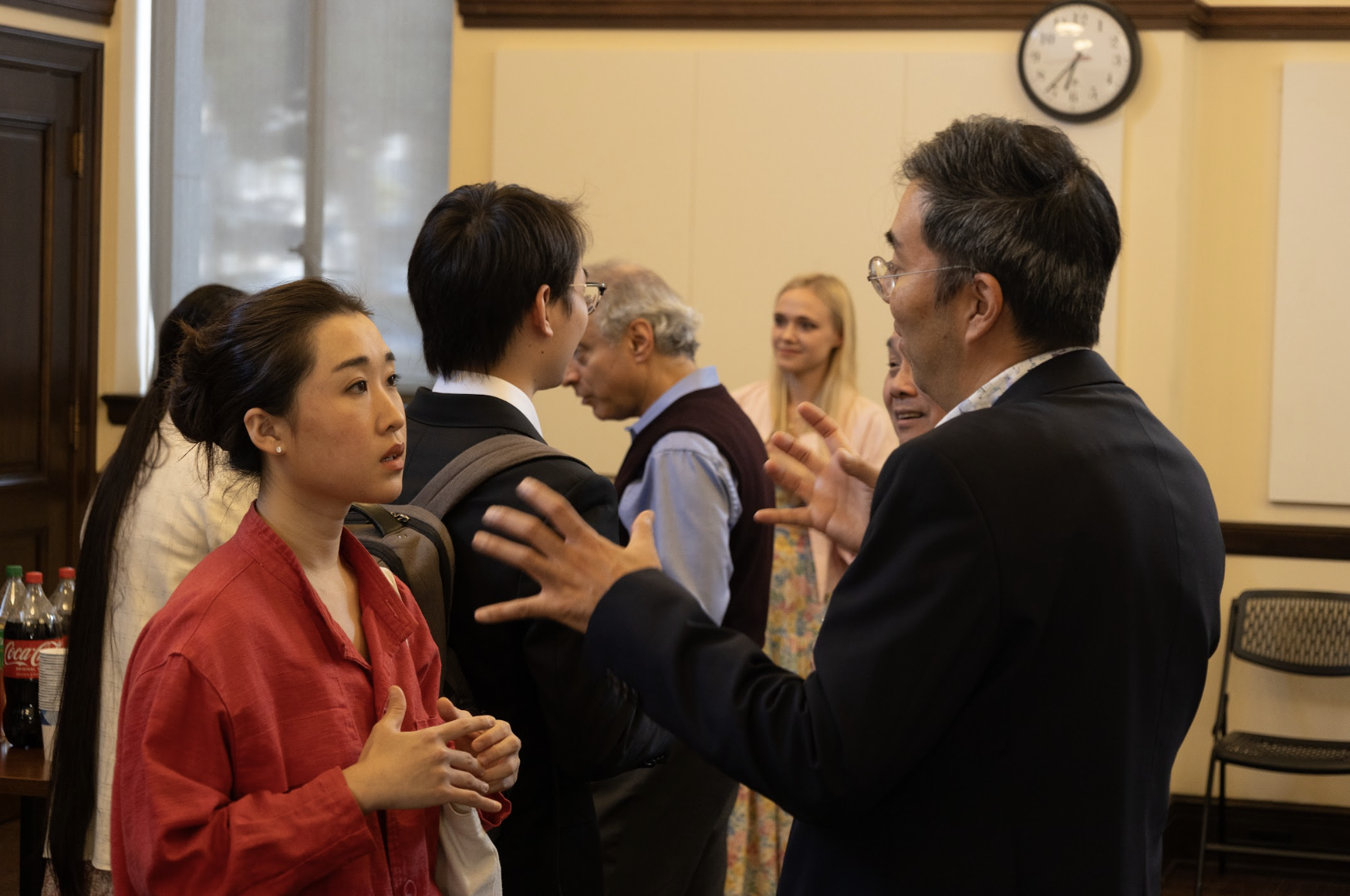 Man speaking to a group of young adults in a room with a clock on the wall.