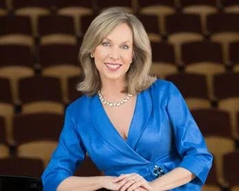A smiling woman with shoulder-length blonde hair wearing a blue blouse and pearl necklace, sitting in what appears to be an auditorium with empty seats in the background.
