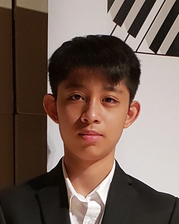 A young man in a black suit with crossed arms, standing in front of a blurred background with a sign and abstract wall art.