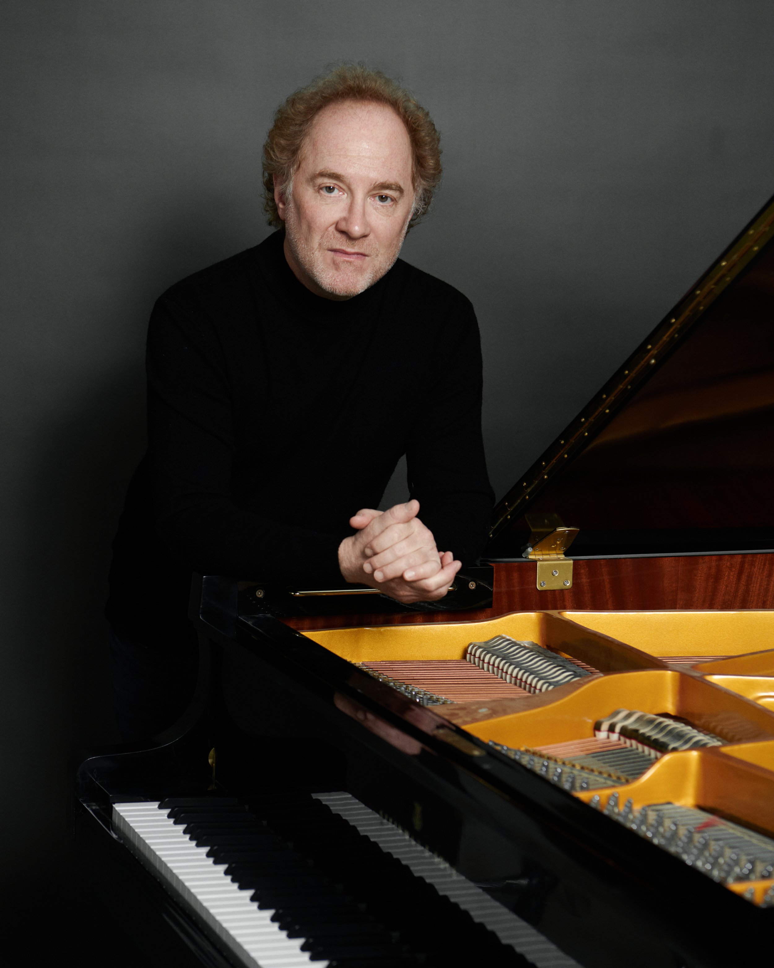A man with curly red hair and a slight smile, wearing a black jacket over a white shirt, sitting in front of a piano.