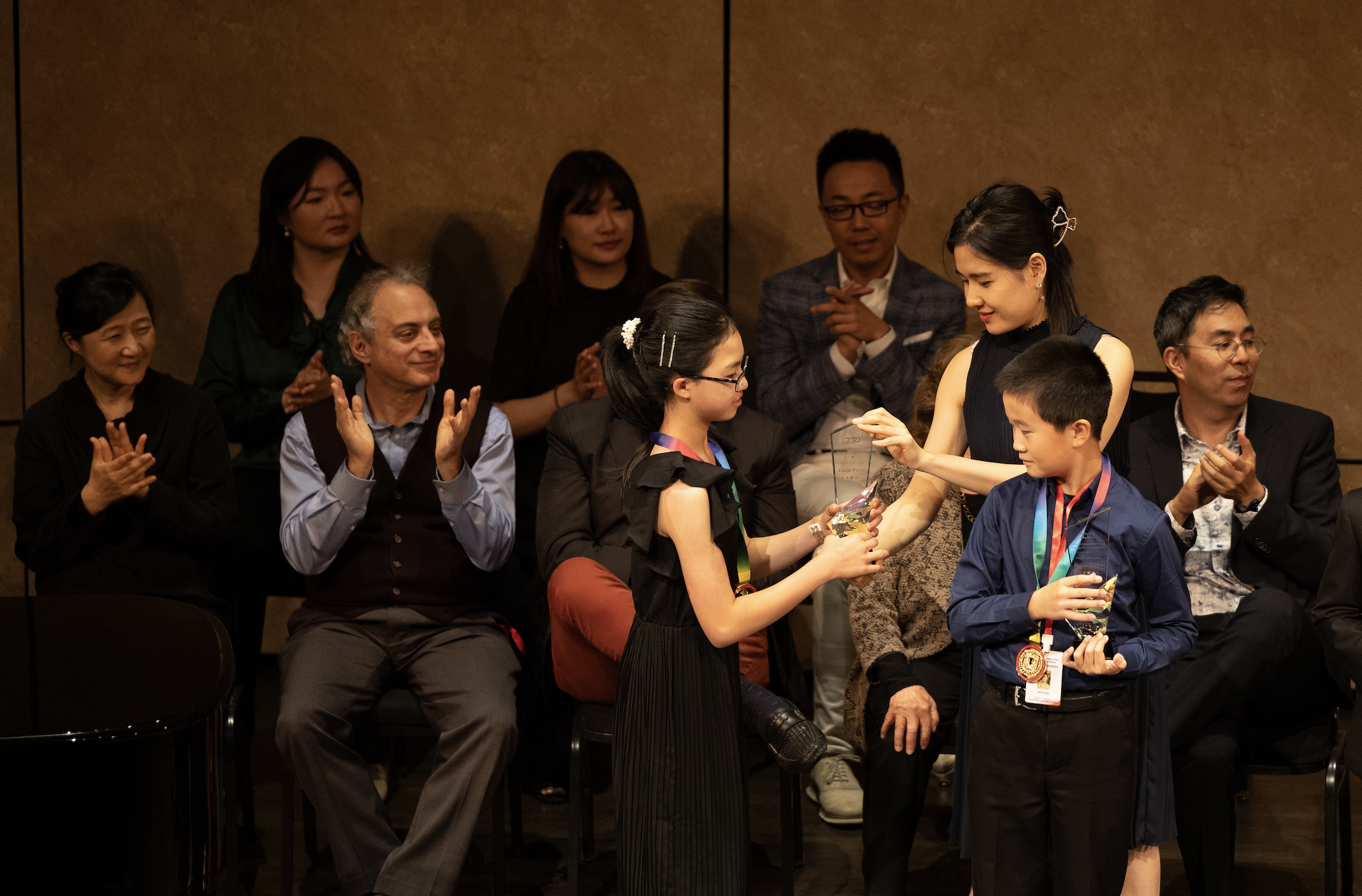 A ceremony with children receiving awards from a woman, while seated adults and children look on and applaud.