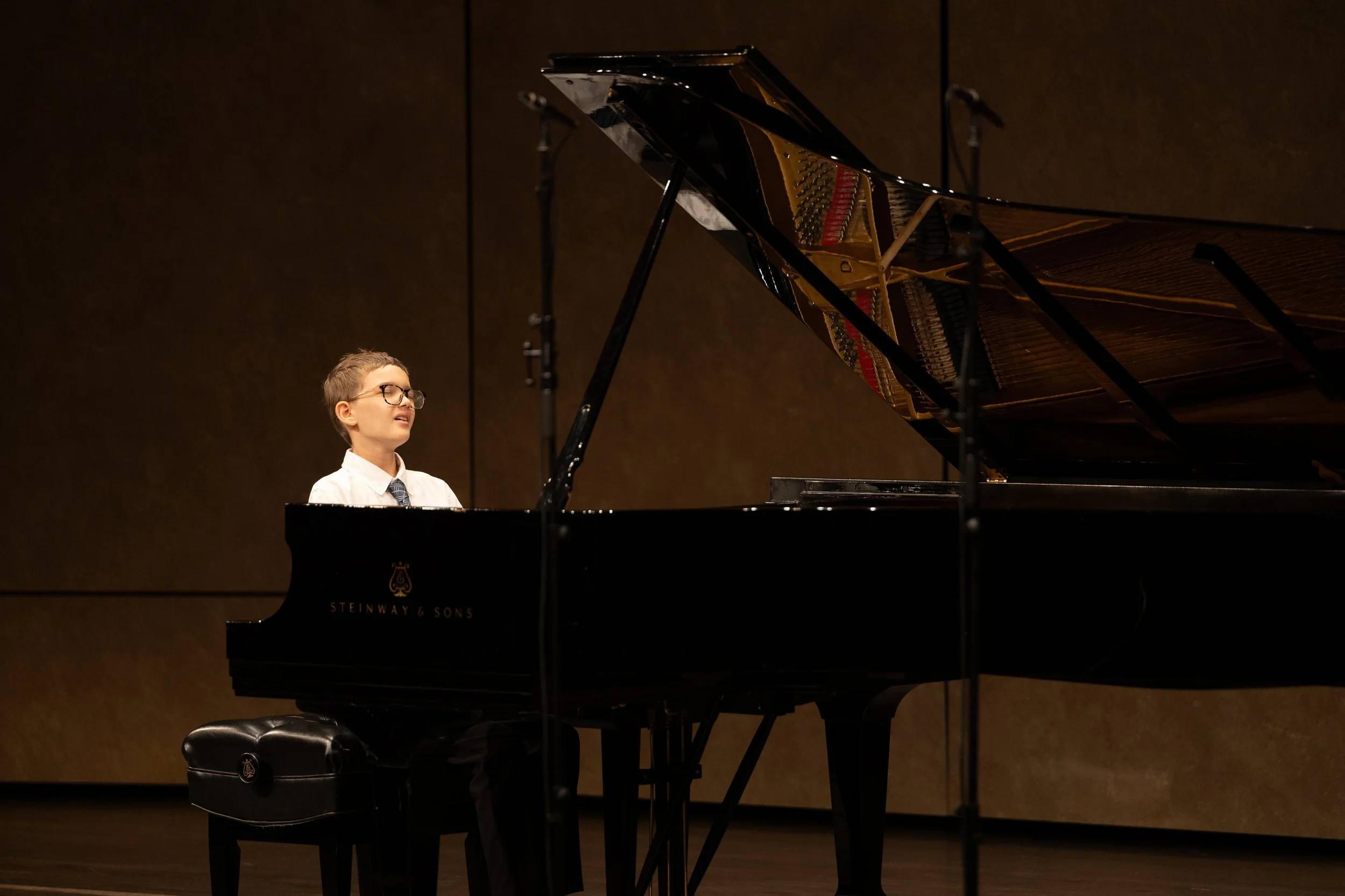 Young boy playing a grand piano on stage with a brown backdrop.