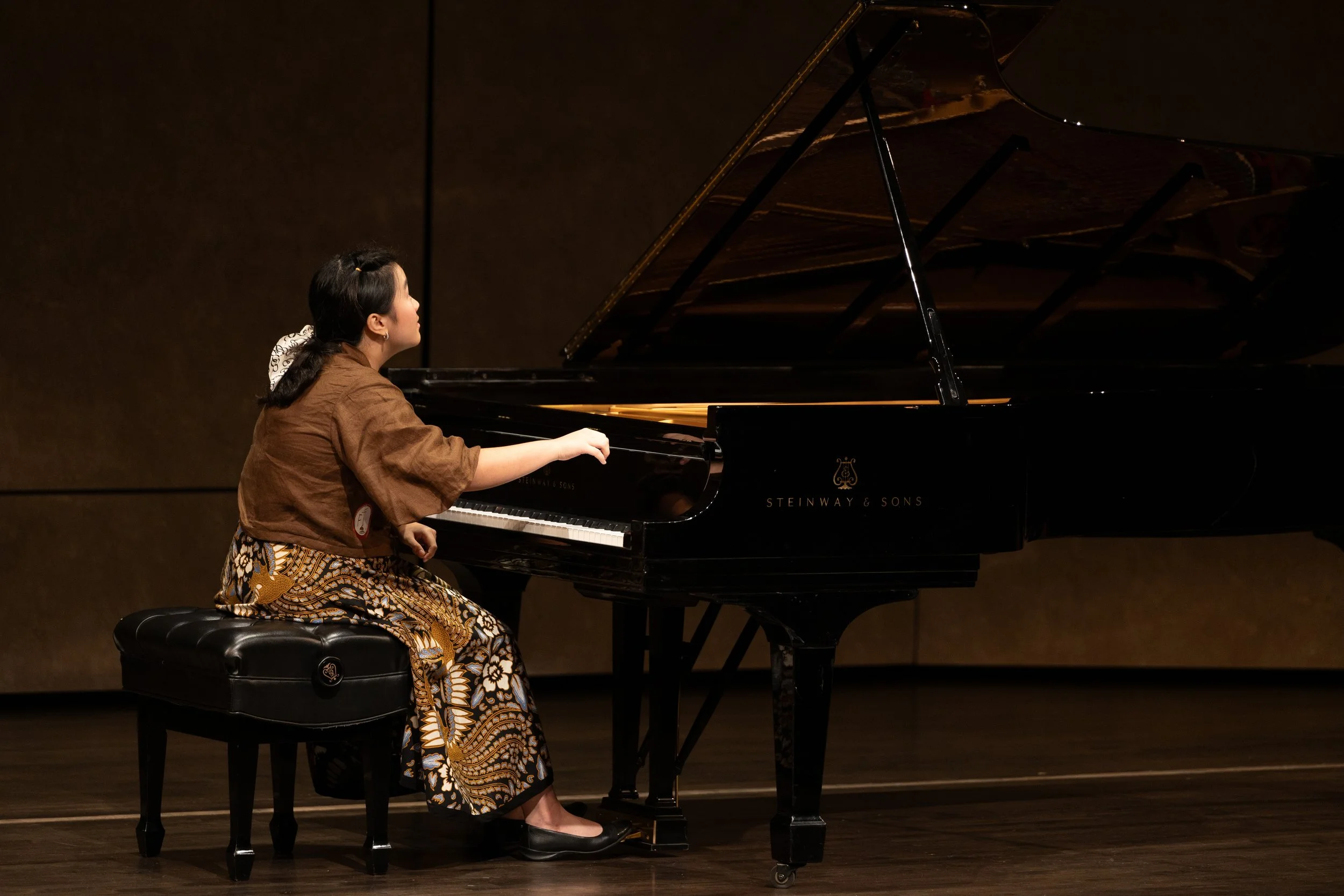 A young girl playing a grand piano on stage, sitting on a black padded stool, in a concert setting.