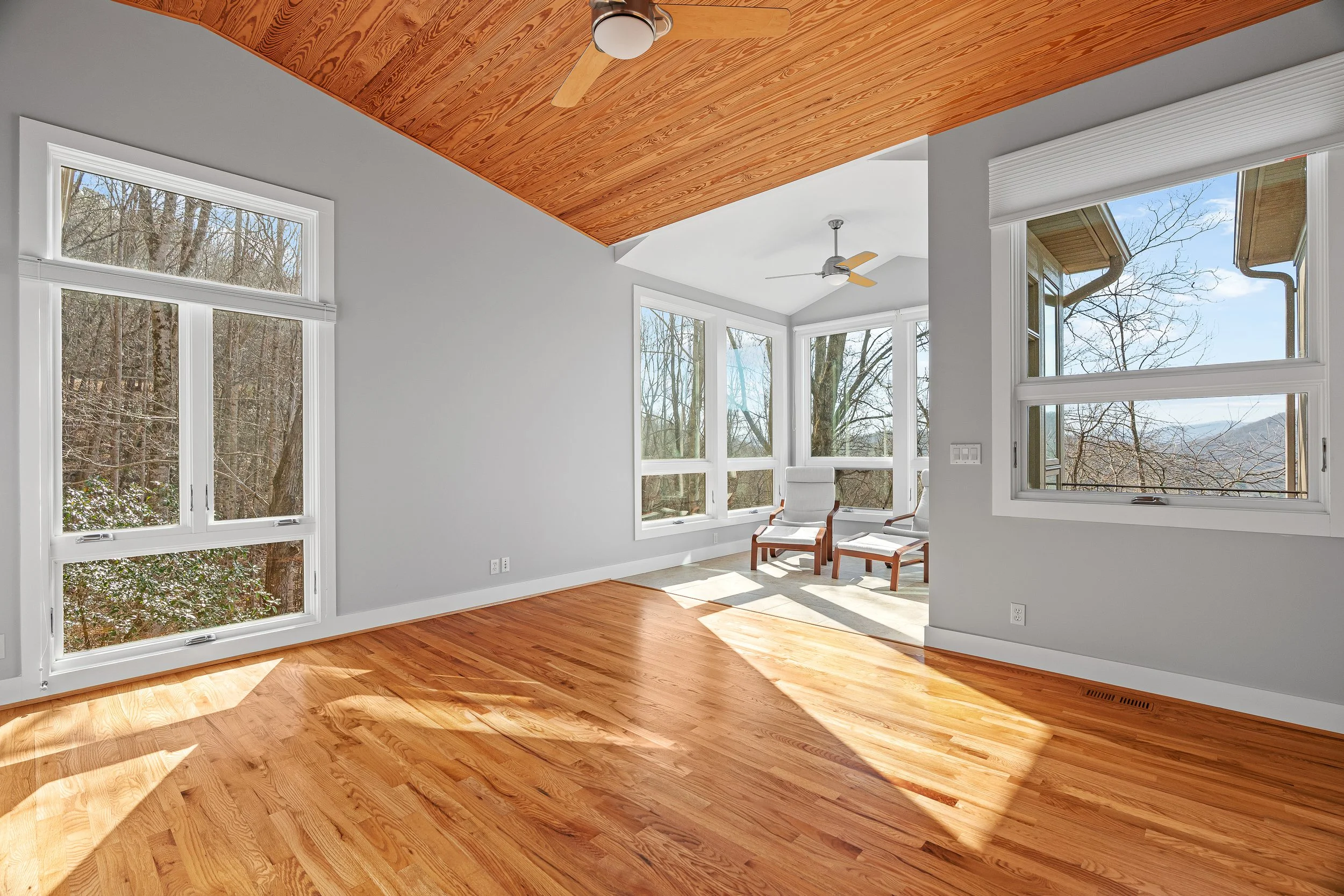 Room with large windows, hardwood floors, white walls, and a wooden ceiling with ceiling fans. Sunlight streams into the room, and a small seating area with two chairs is visible near the windows.