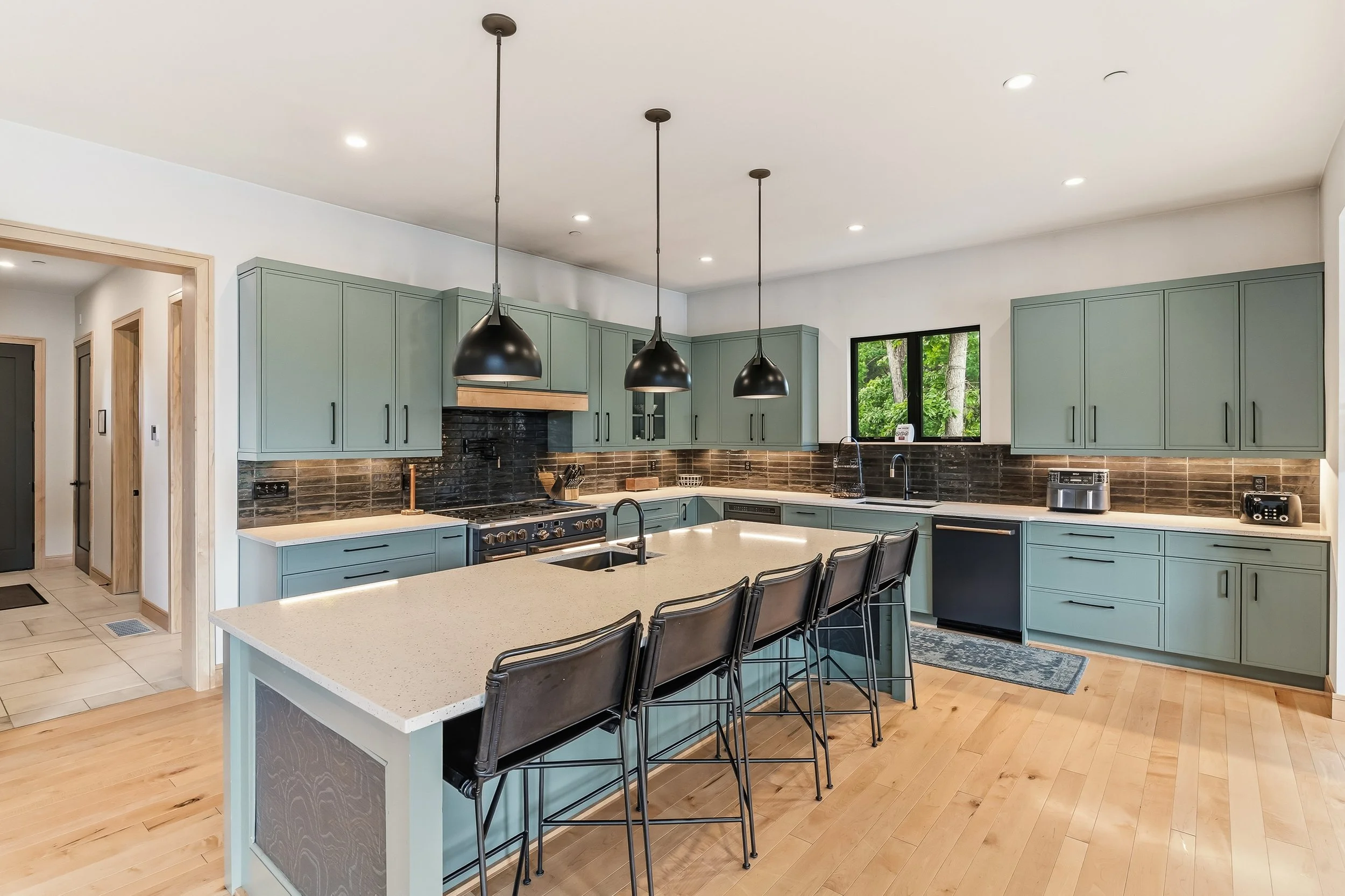 Modern kitchen with mint green cabinets, black pendant lights, large kitchen island, black faucet, window with view of trees, black and stainless appliances, hardwood floors, and wood trim around doorways.