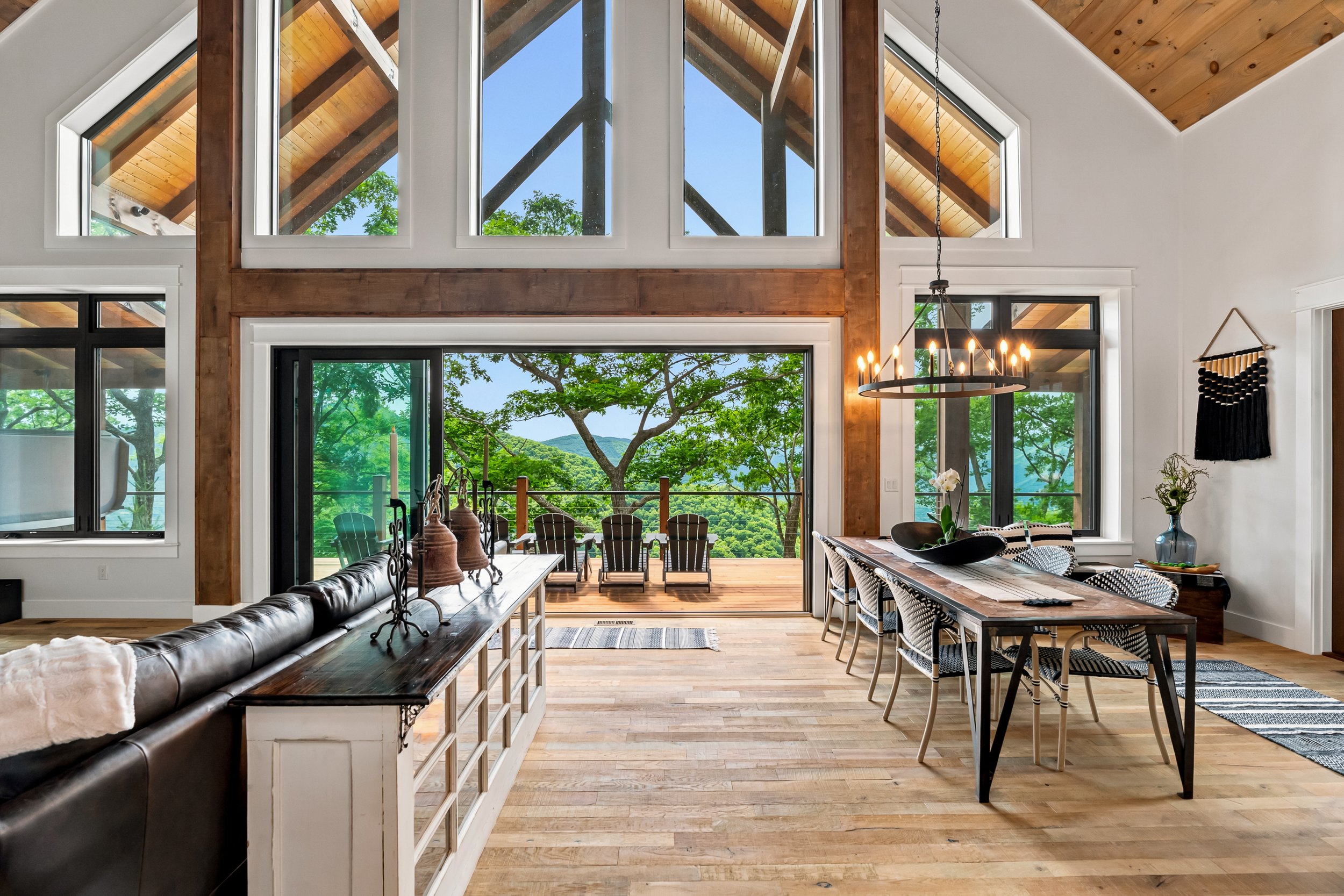 Interior of a living space with large windows and a view of trees and mountains outside. There is a wooden dining table with chairs, a black leather sofa, decorative items, and a view of an outdoor balcony with chairs.