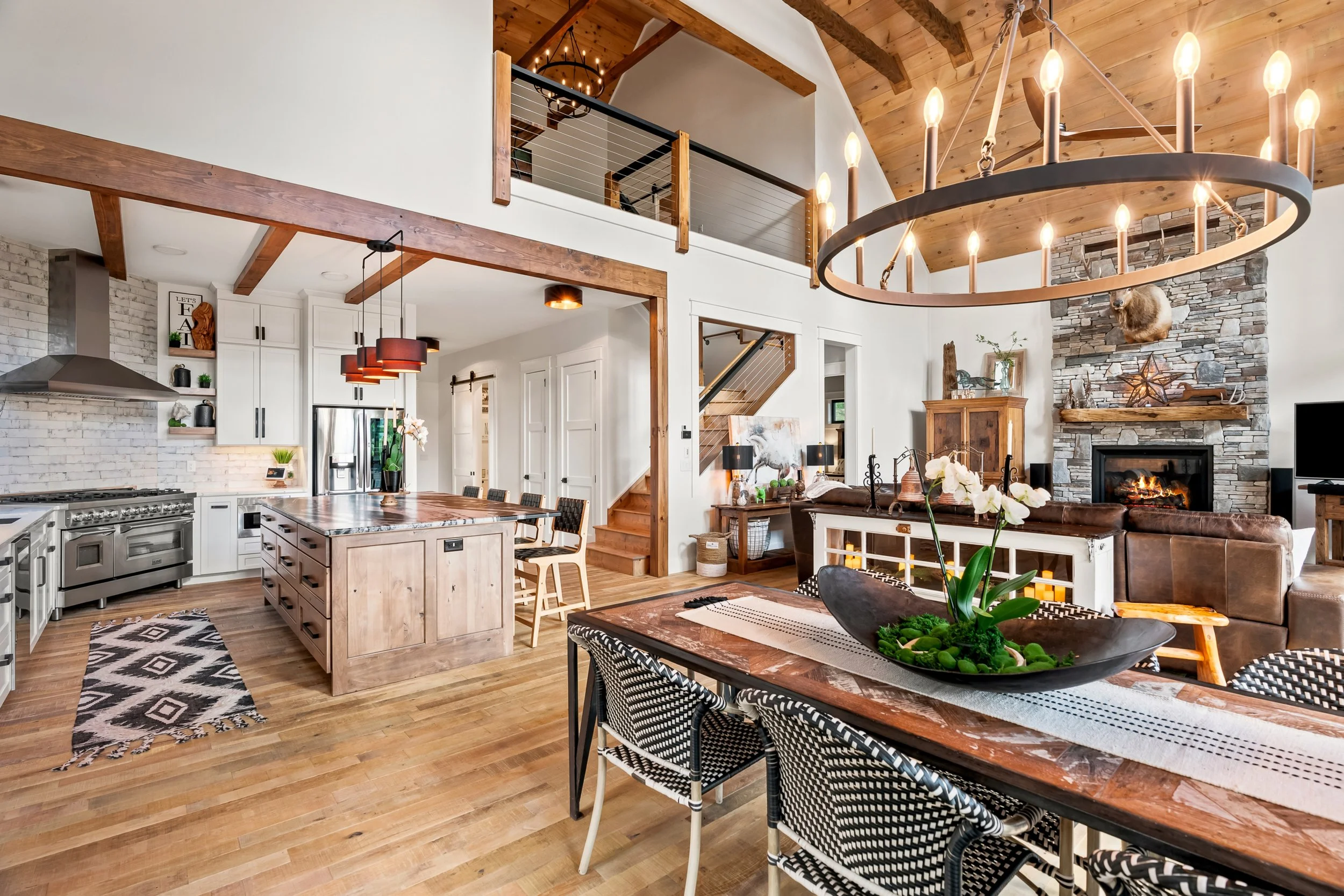 Open concept living room and kitchen with wood accents, a large round chandelier with candle-like lights, a stone fireplace with a mounted animal skull, and a wooden staircase leading to an upper level.