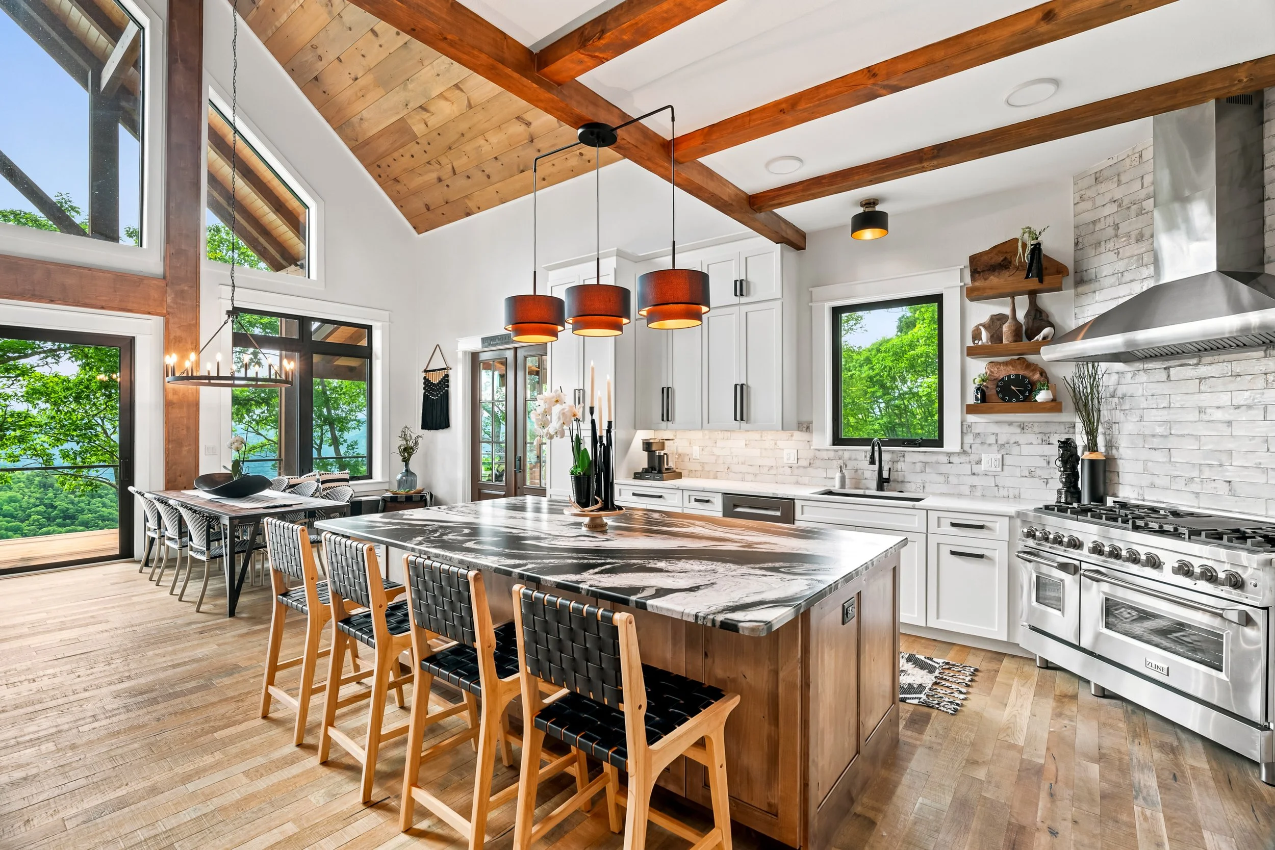 Modern kitchen and dining area with large windows showing greenery outside, white cabinets, a marble kitchen island with wooden chairs, a stainless steel stove, and decorative wall shelves with plants and art.