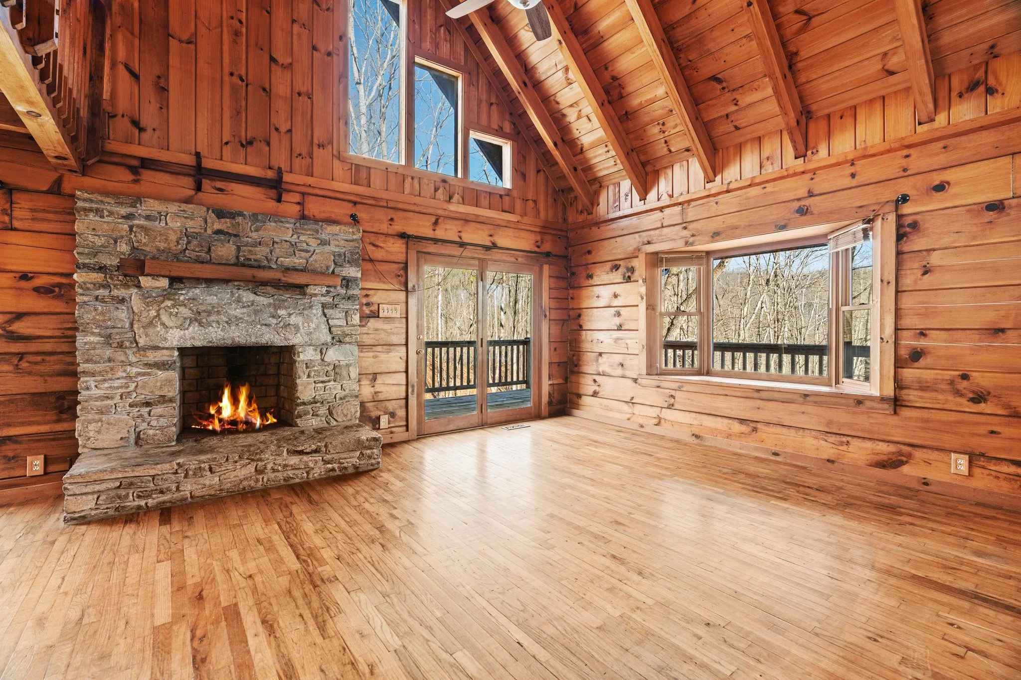 Empty living room with wooden walls, ceiling, and floor, fireplace with a fire, large windows, and sliding glass door leading outside.
