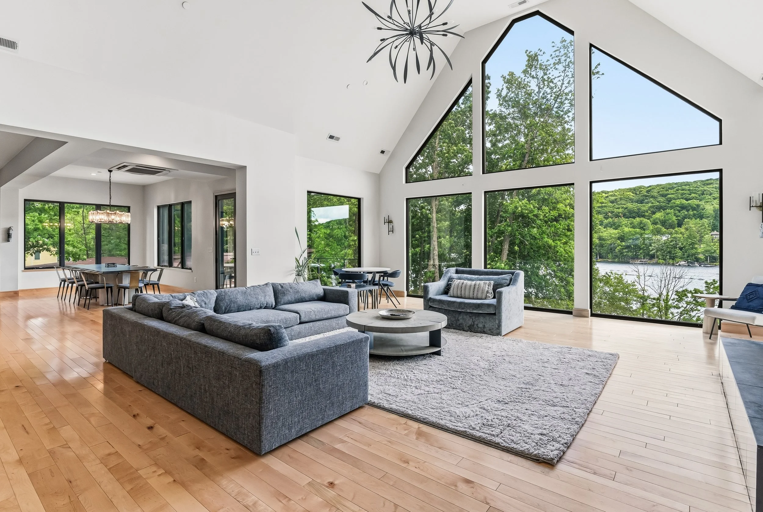 Interior of a modern living room with large floor-to-ceiling windows showing trees and a water view outside. The room has a gray sofa, armchair, round coffee table, and wooden flooring. There is a dining area with a table and chairs, and a contempora