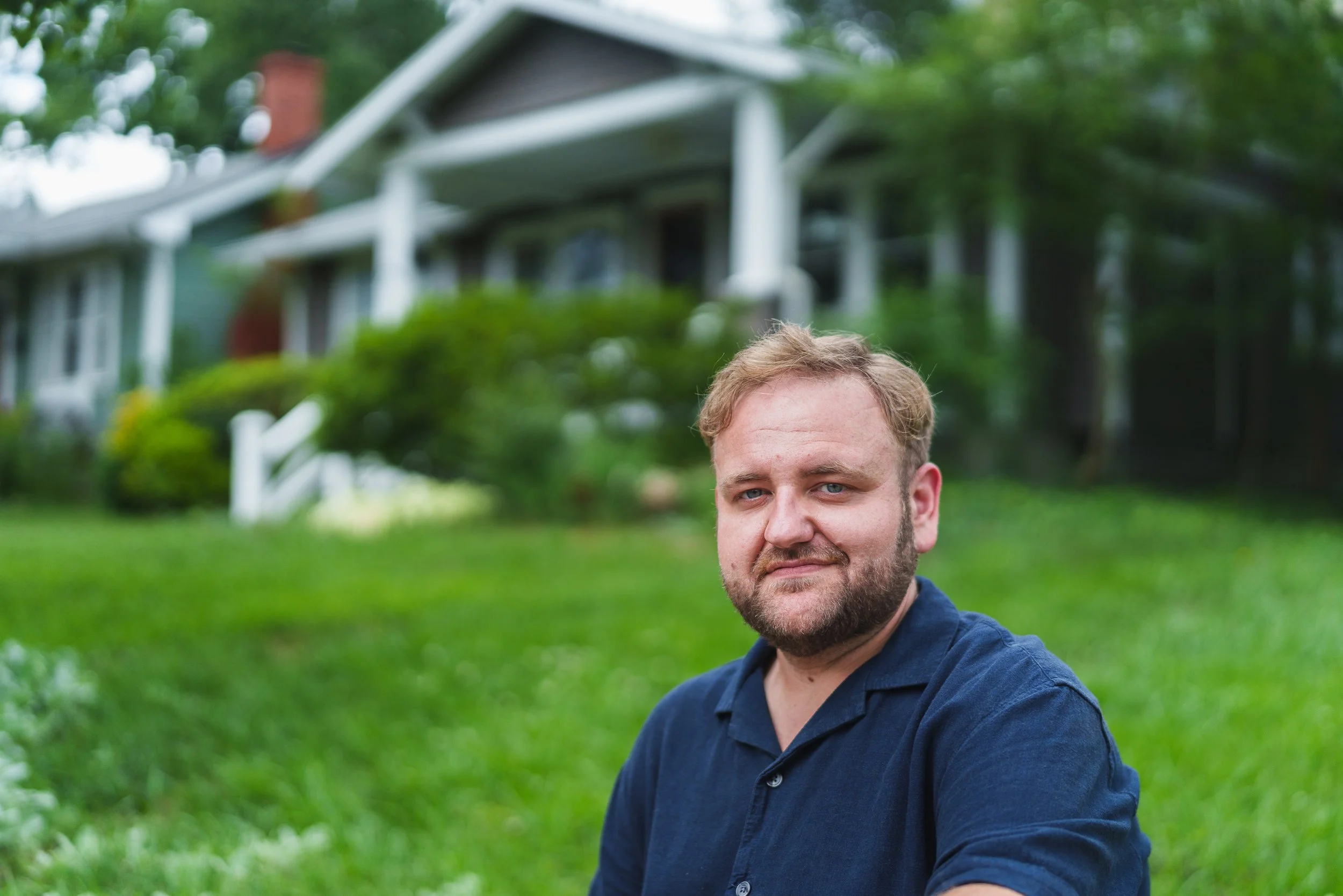 A man with light brown hair and a beard, wearing a navy blue shirt, sitting outdoors on grass with house and trees in the background.
