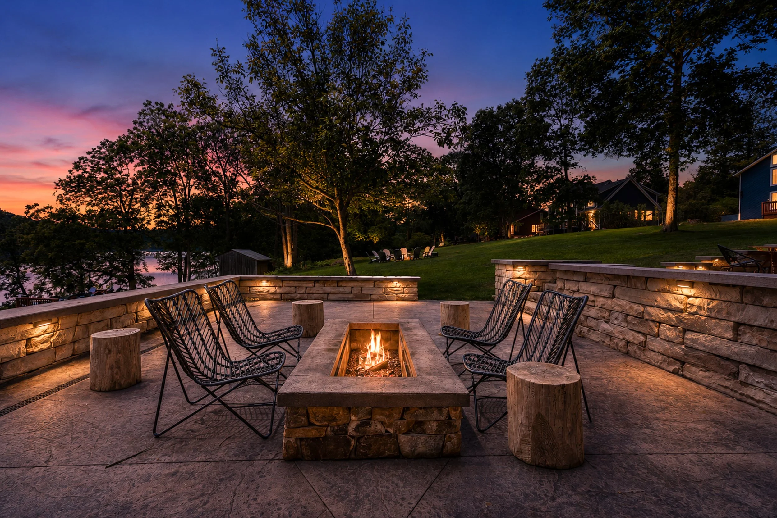 Outdoor patio with a stone fire pit surrounded by black chairs, overlooking trees and houses at sunset with a colorful sky.