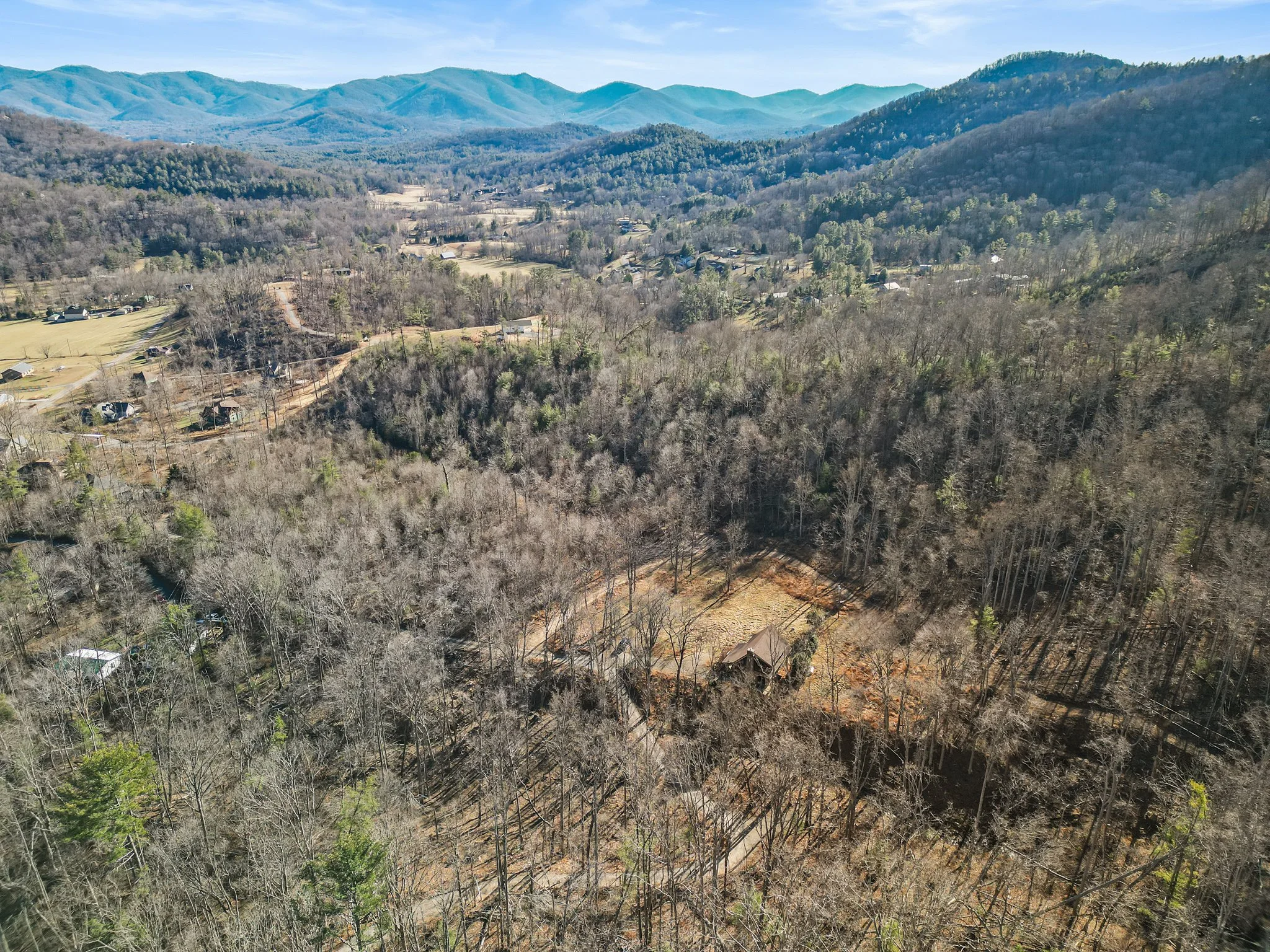 Aerial view of a wooded mountain landscape with a small house and a clearing, surrounded by barren trees and distant mountains in the background.