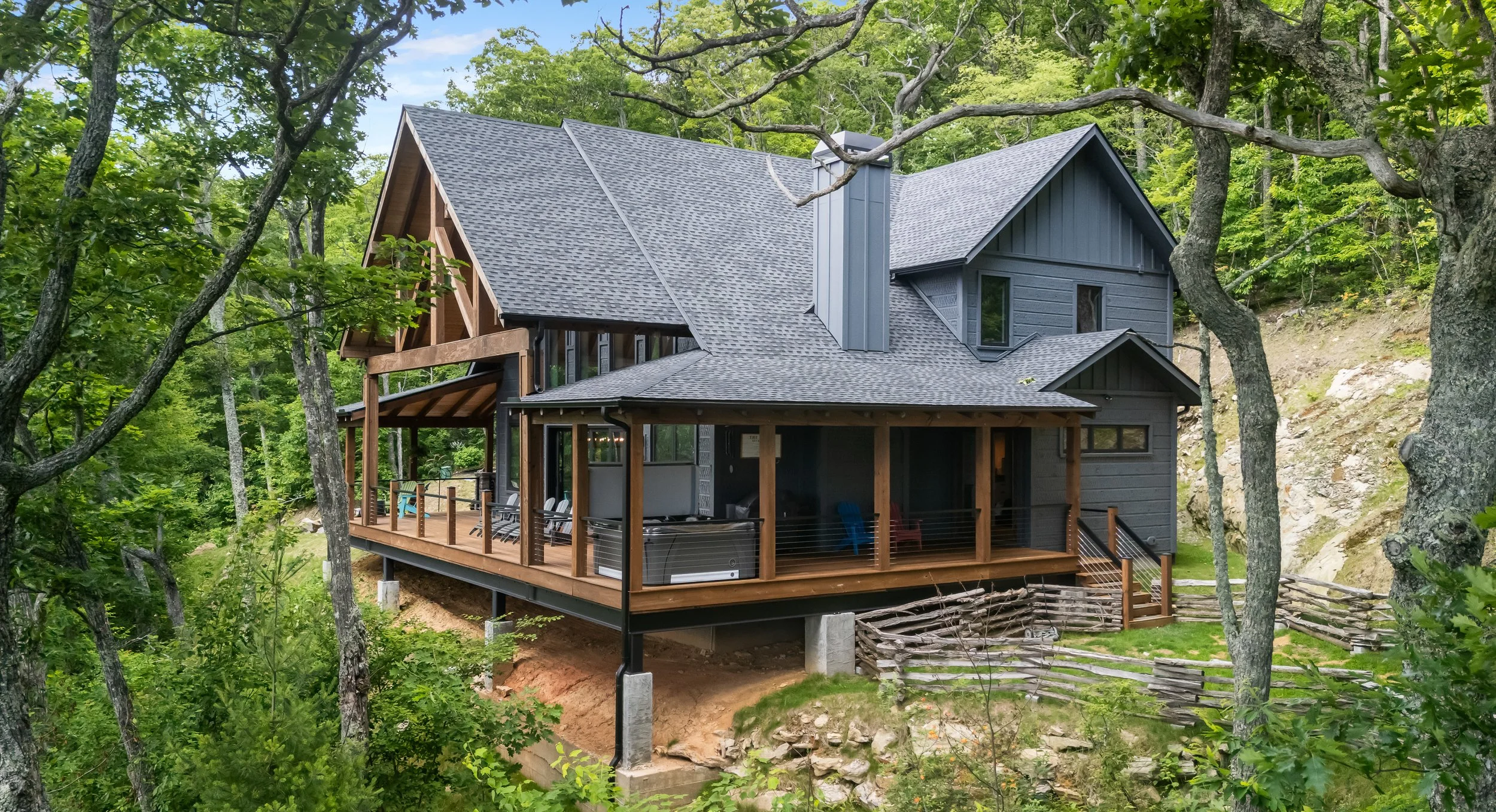 A modern house with gray siding and a steeply pitched roof, surrounded by lush green trees, with a wooden deck and outdoor furniture.