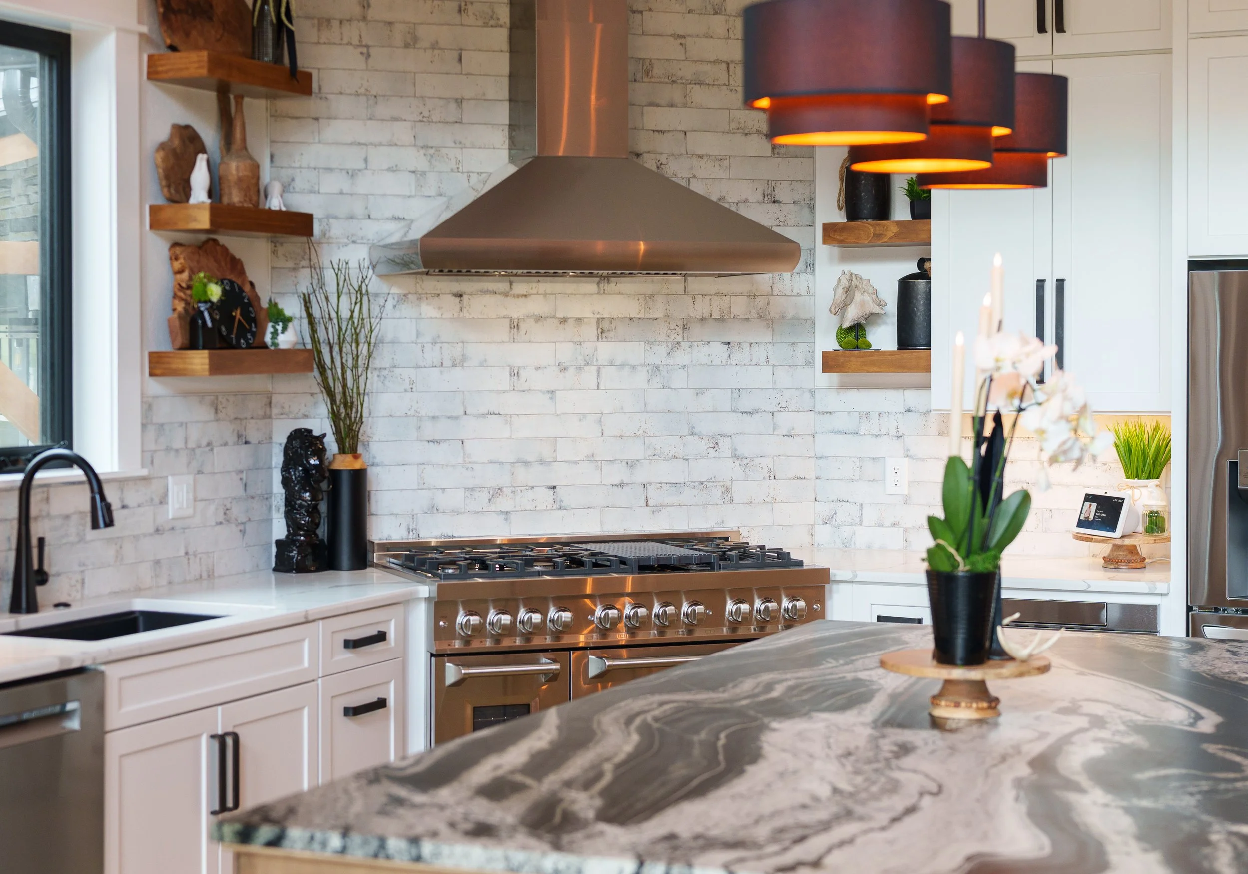 Modern kitchen with white cabinets, marble countertops, a stainless steel gas stove, and a white brick wall with open shelves holding decorative items and plants. Pendant lights hang above the counter.