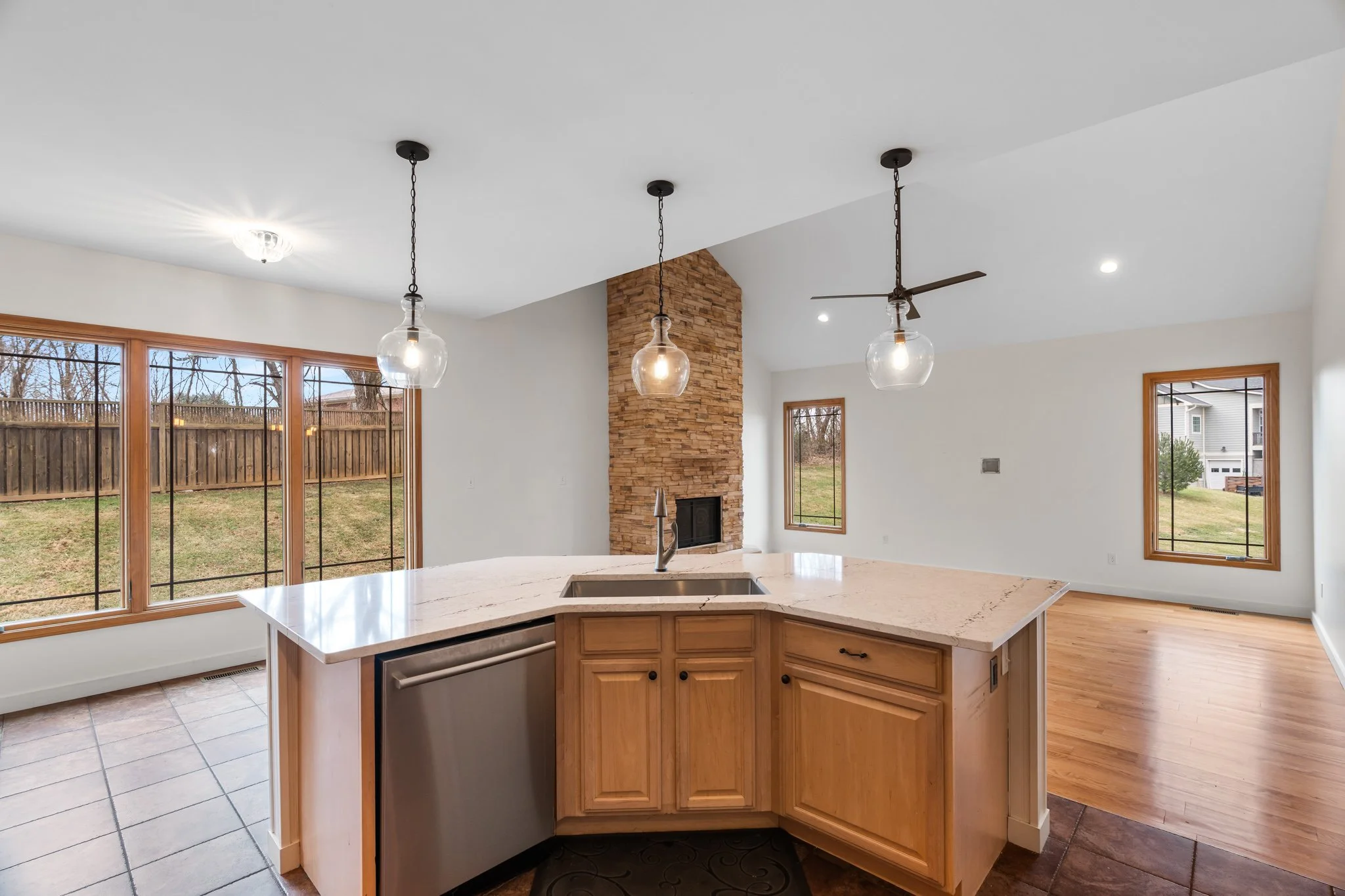 Open kitchen and living room area with large windows, stone fireplace, kitchen island with marble countertop, wooden cabinets, pendant lights, ceiling fan, hardwood and tile flooring.