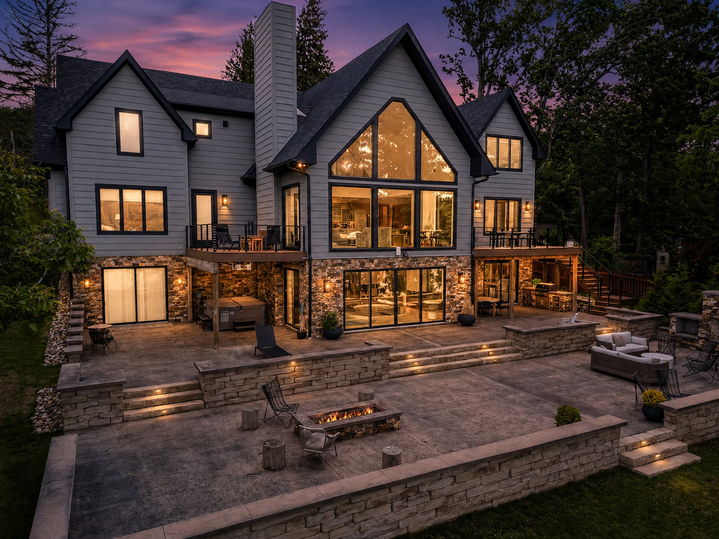 A large, modern house with gray siding, multiple gabled roofs, and large windows illuminated from inside. The house is situated in a wooded area at dusk, with the sky showing purple and pink hues. The backyard features a stone patio, outdoor seating,