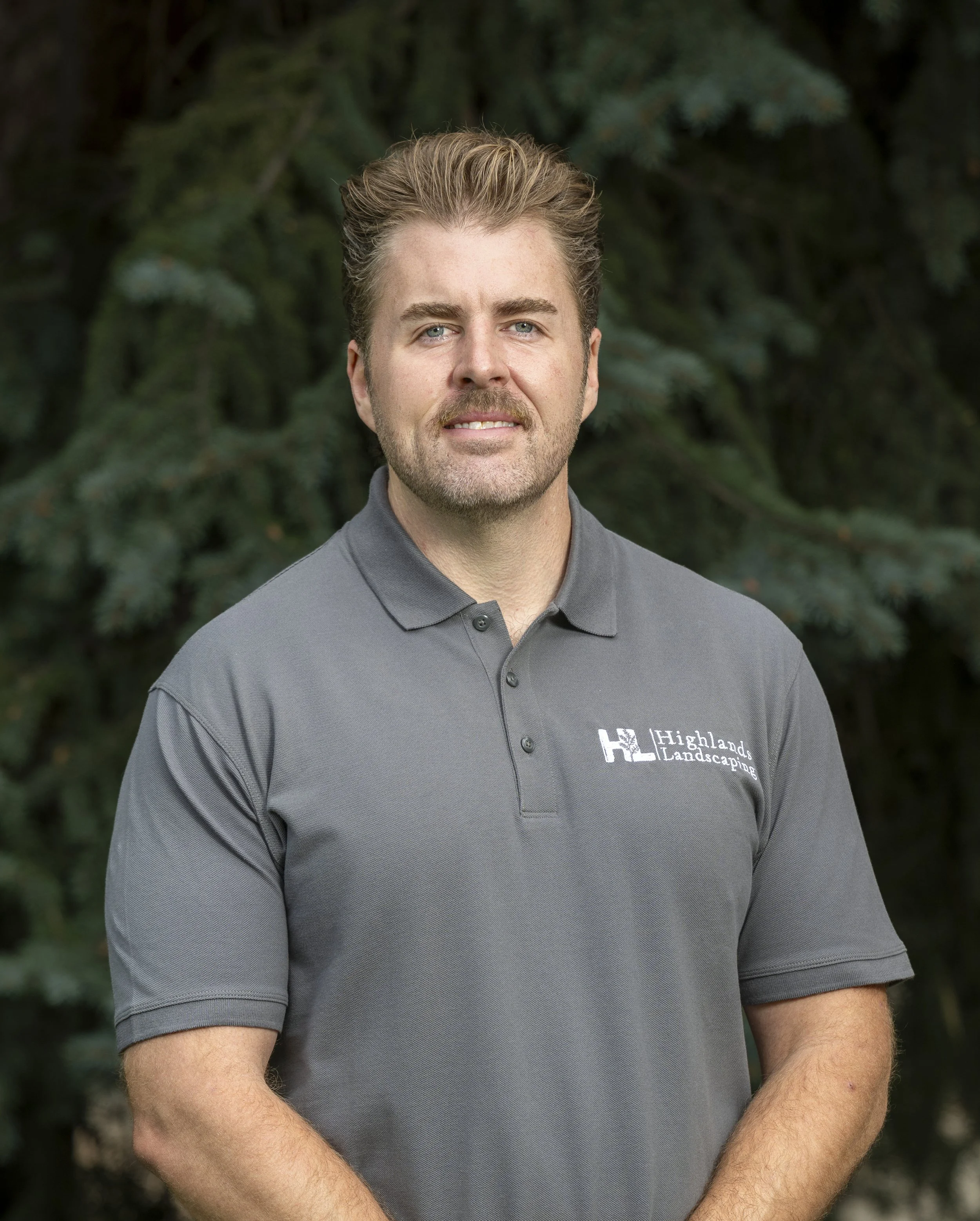 Young man wearing a Highlands Landscaping cap and polo shirt stands outdoors in front of green foliage, looking at the camera.