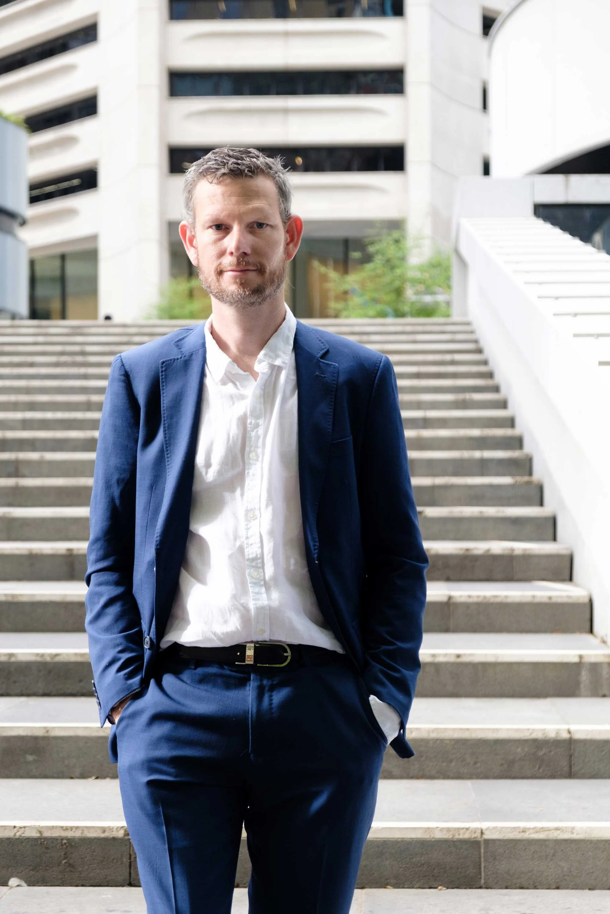 Justin Barnes Principal Mortgage Broker and Managing Director of Barco Consulting Group home of Barco Consulting and Barco Finance in blue suit and white shirt standing on outdoor steps, modern building in background.