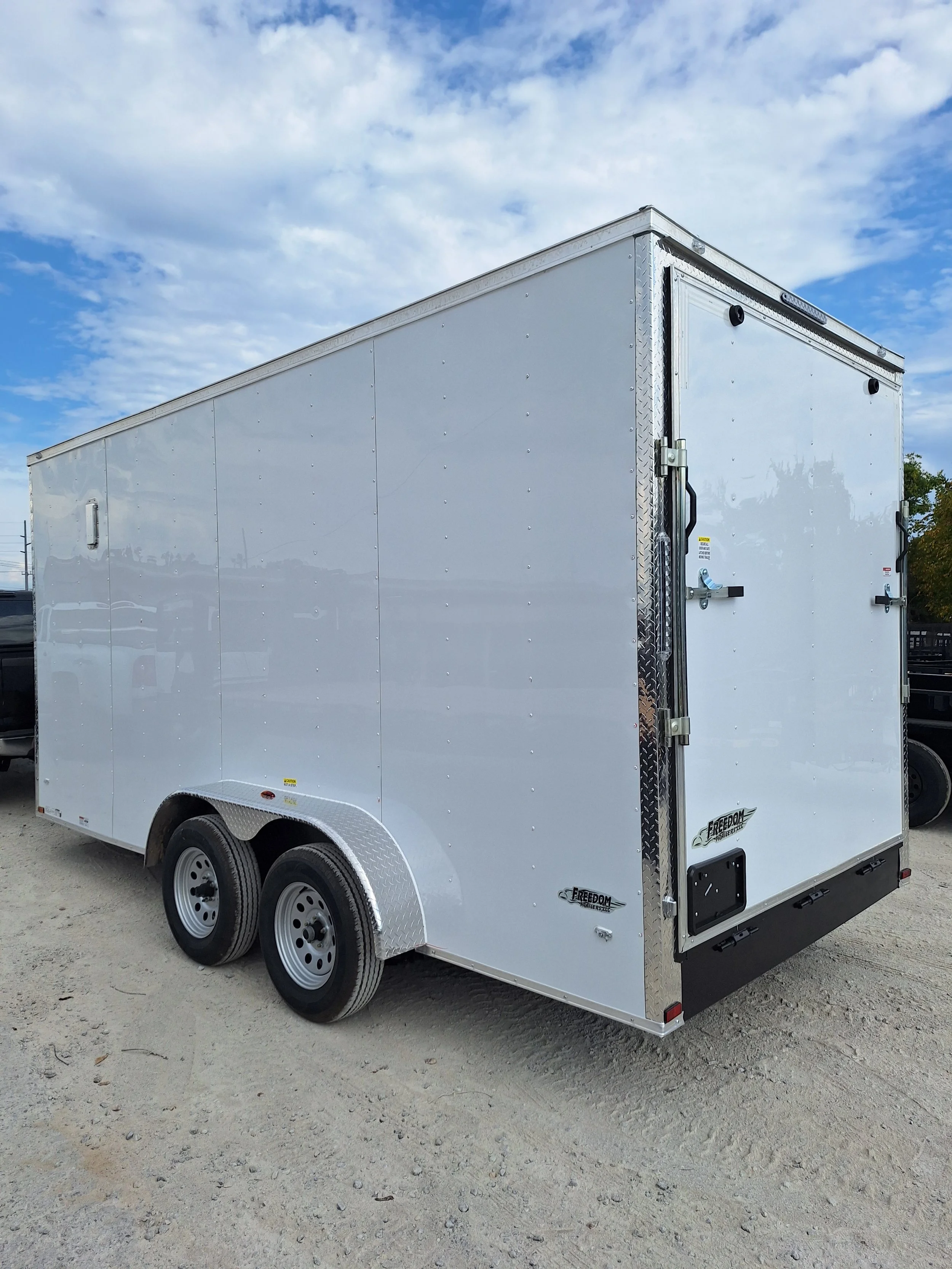 White enclosed cargo trailer with dual axles parked on a dirt lot under a partly cloudy sky.