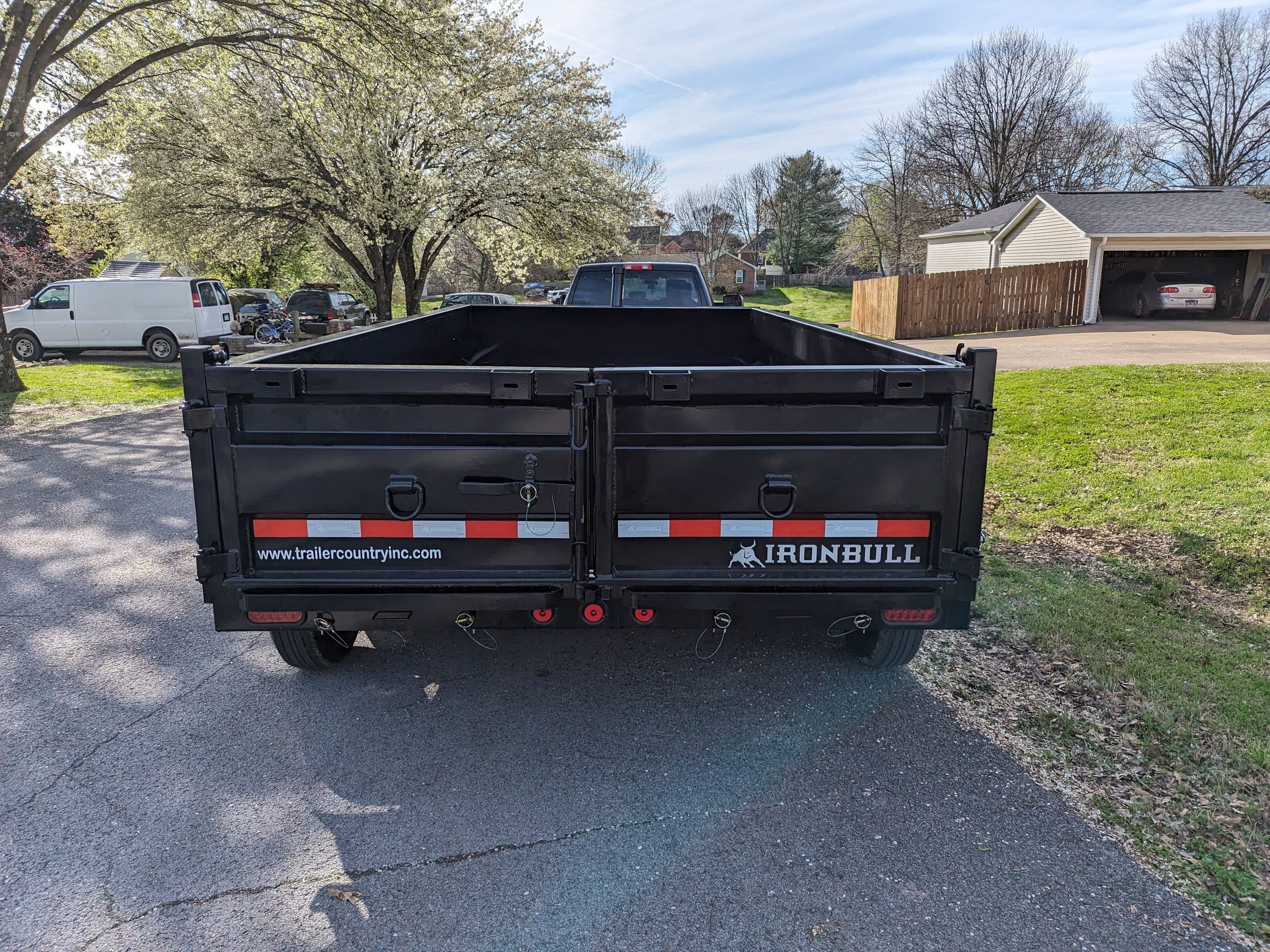 Black iron trailer parked on a residential street, branded with "IRON BULL" and "trailercountryinc.com", surrounded by trees and homes.