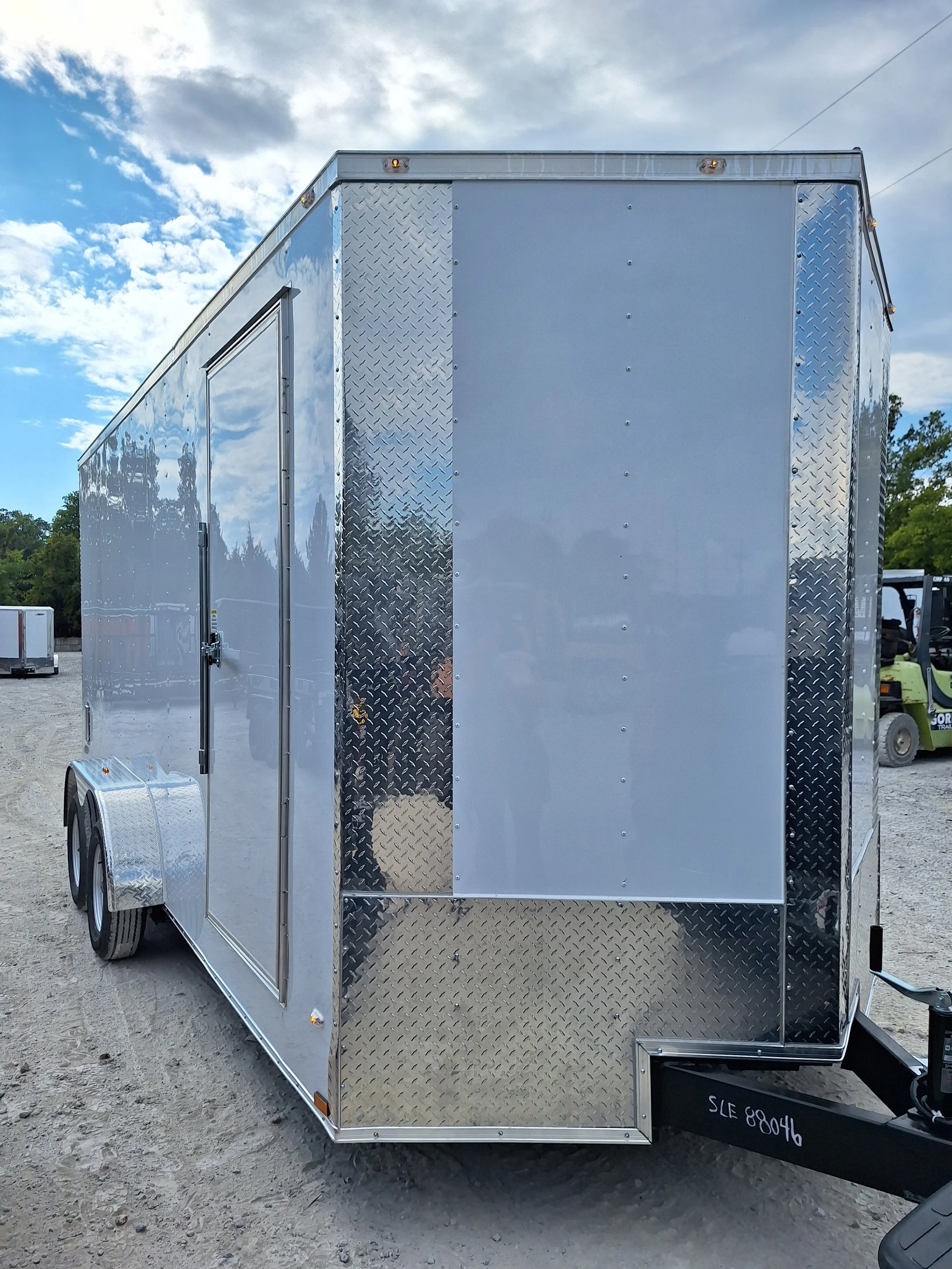 Silver enclosed cargo trailer with diamond plate accents, parked on gravel with a cloudy sky and trees in the background.