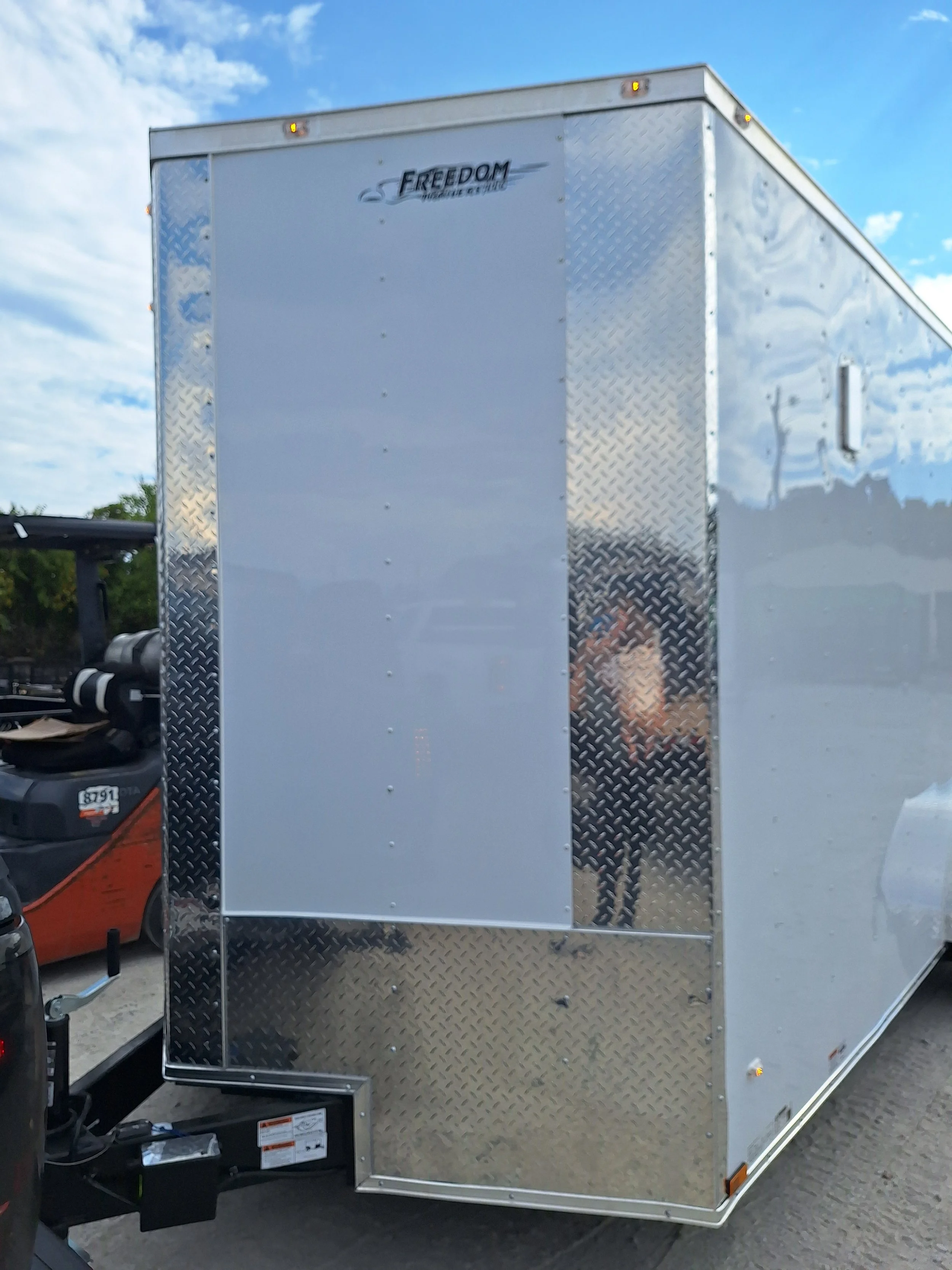 A white enclosed cargo trailer with a diamond plate front, parked on a gravel surface, with a person visible through the reflection taking a photo.