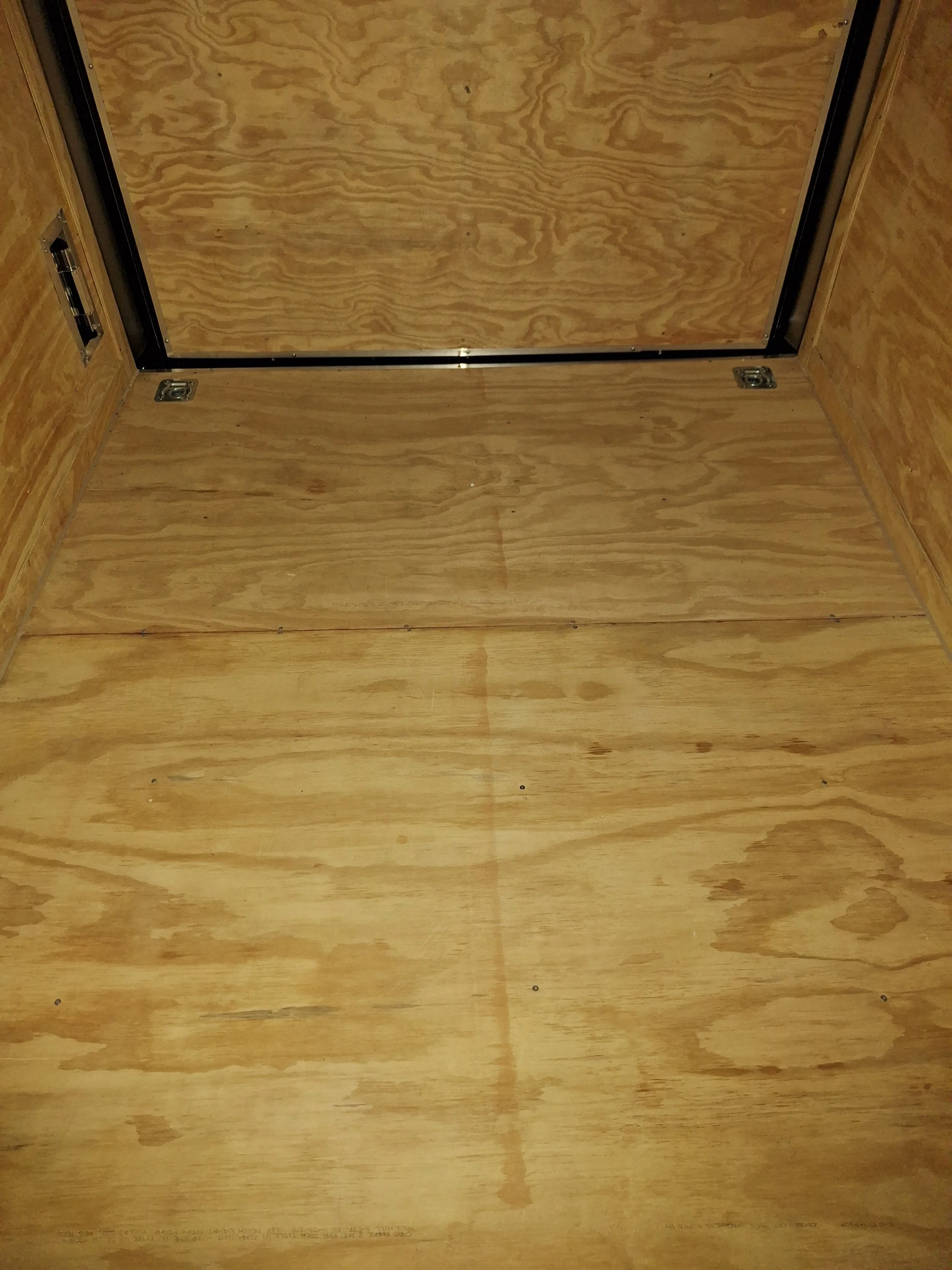 Wooden interior of a small elevator or lift with a plywood floor and walls, and a closed wooden door at the top with a metal frame.