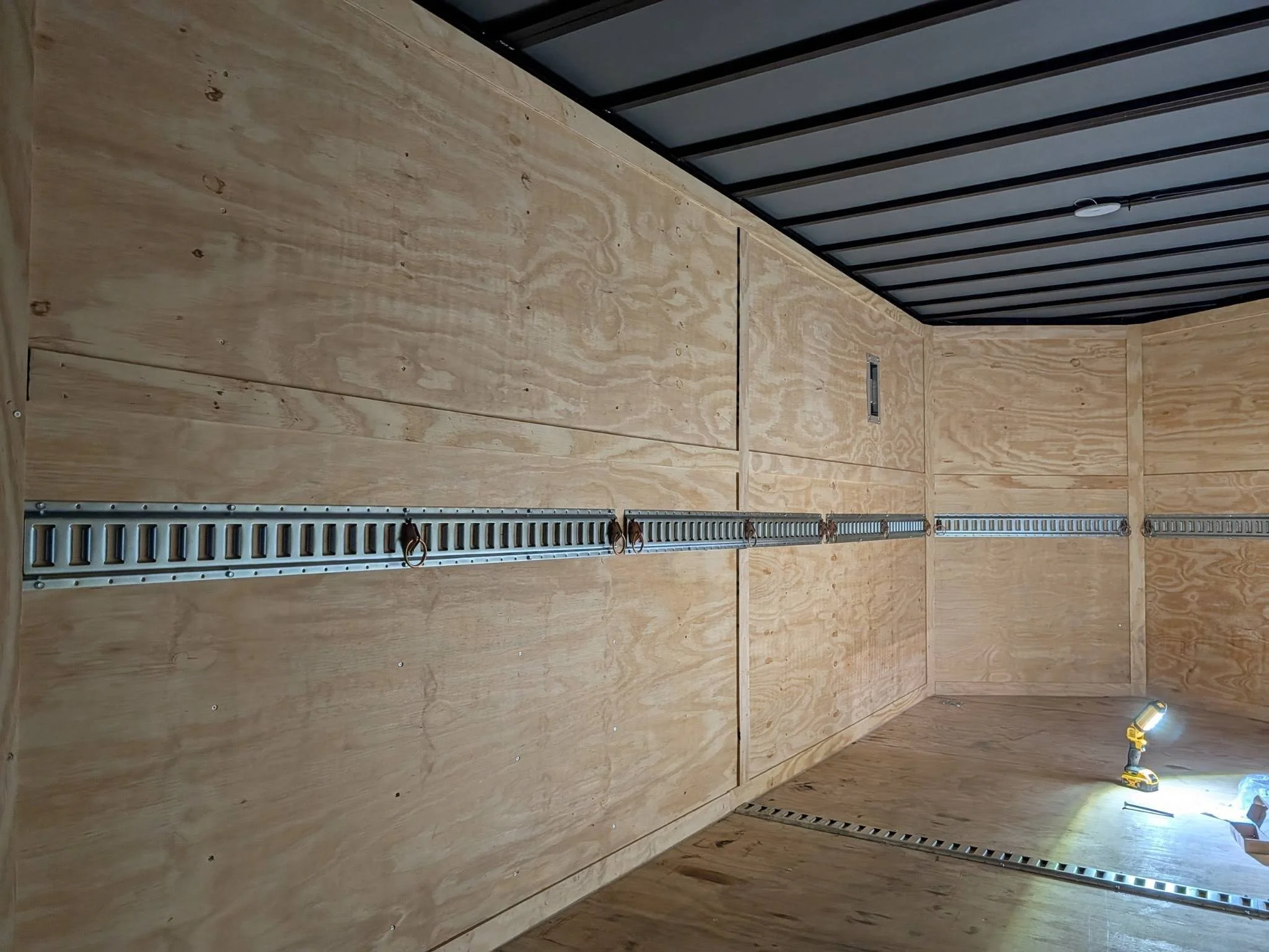 Interior of an unfinished wooden trailer or storage space with plywood walls, a metal track for hanging items, a small wall opening, and a portable work light.