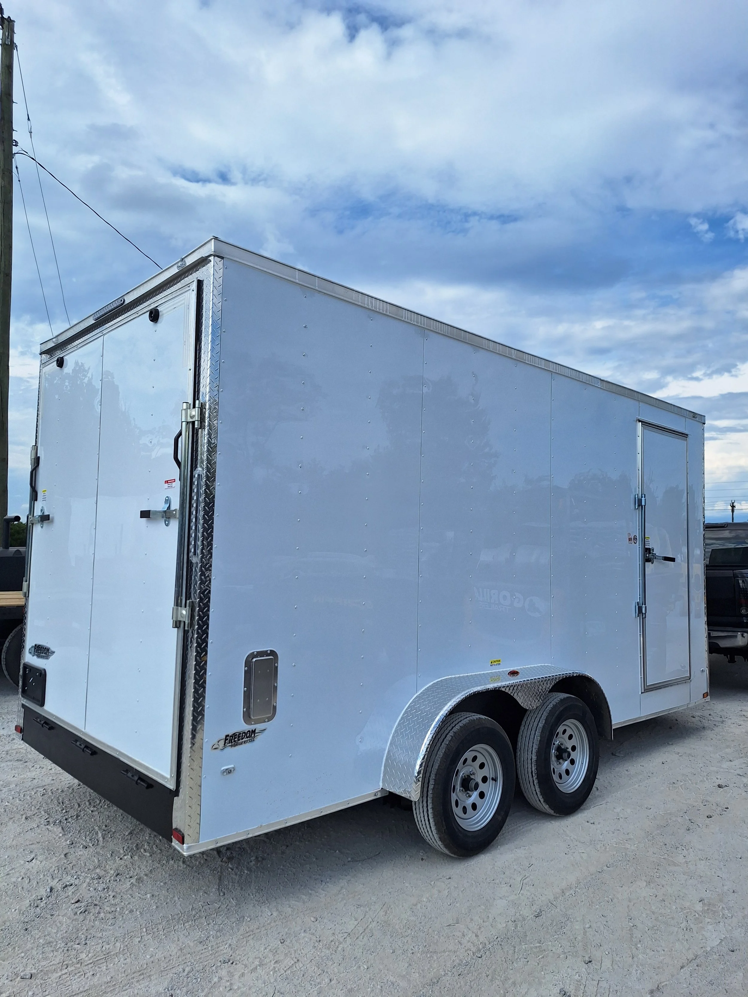 A white enclosed utility trailer with dual axles parked on a gravel lot under a partly cloudy sky.