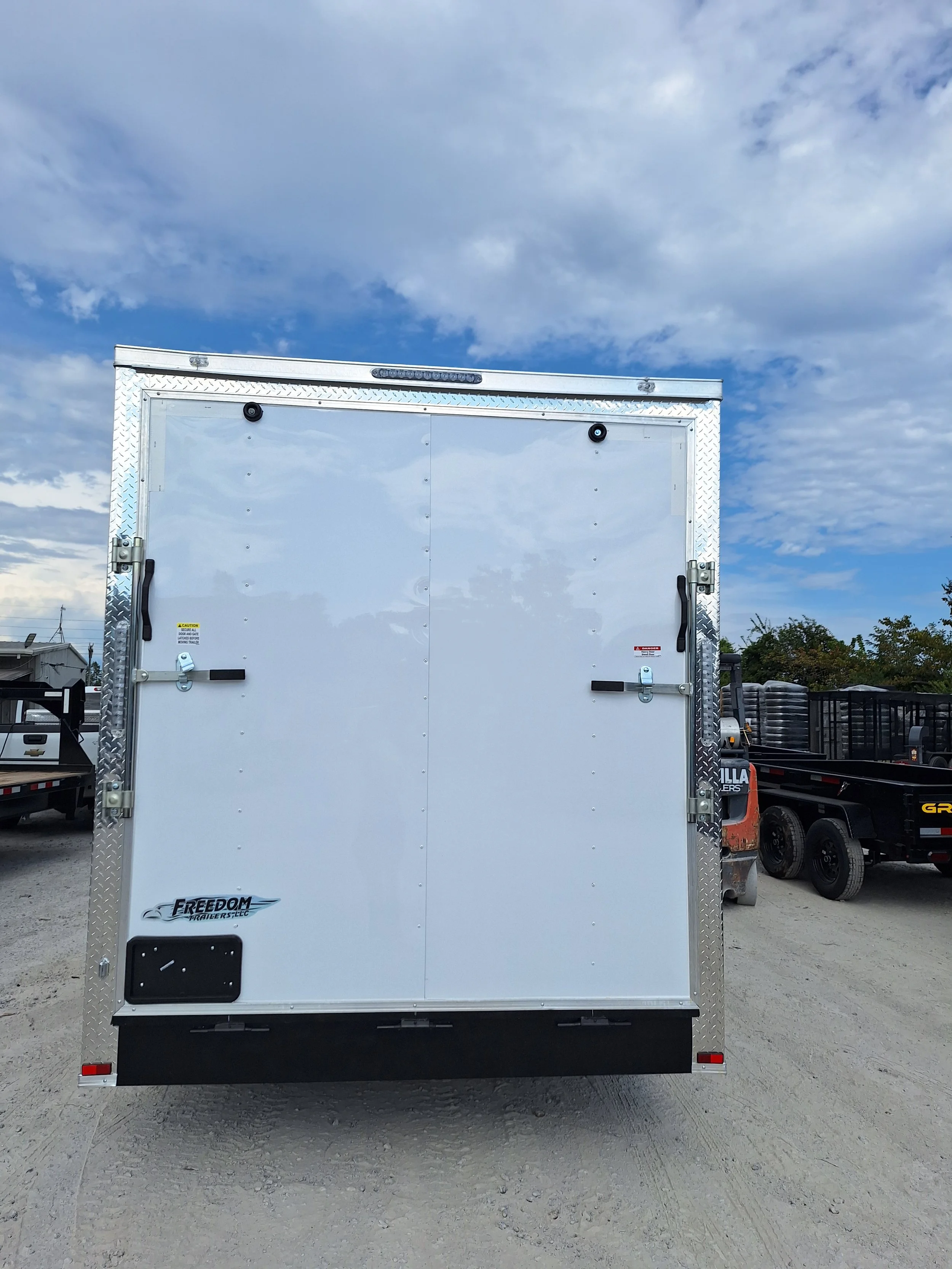 Rear view of a white enclosed cargo trailer with a closed door, parked on a gravel lot under a partly cloudy sky.