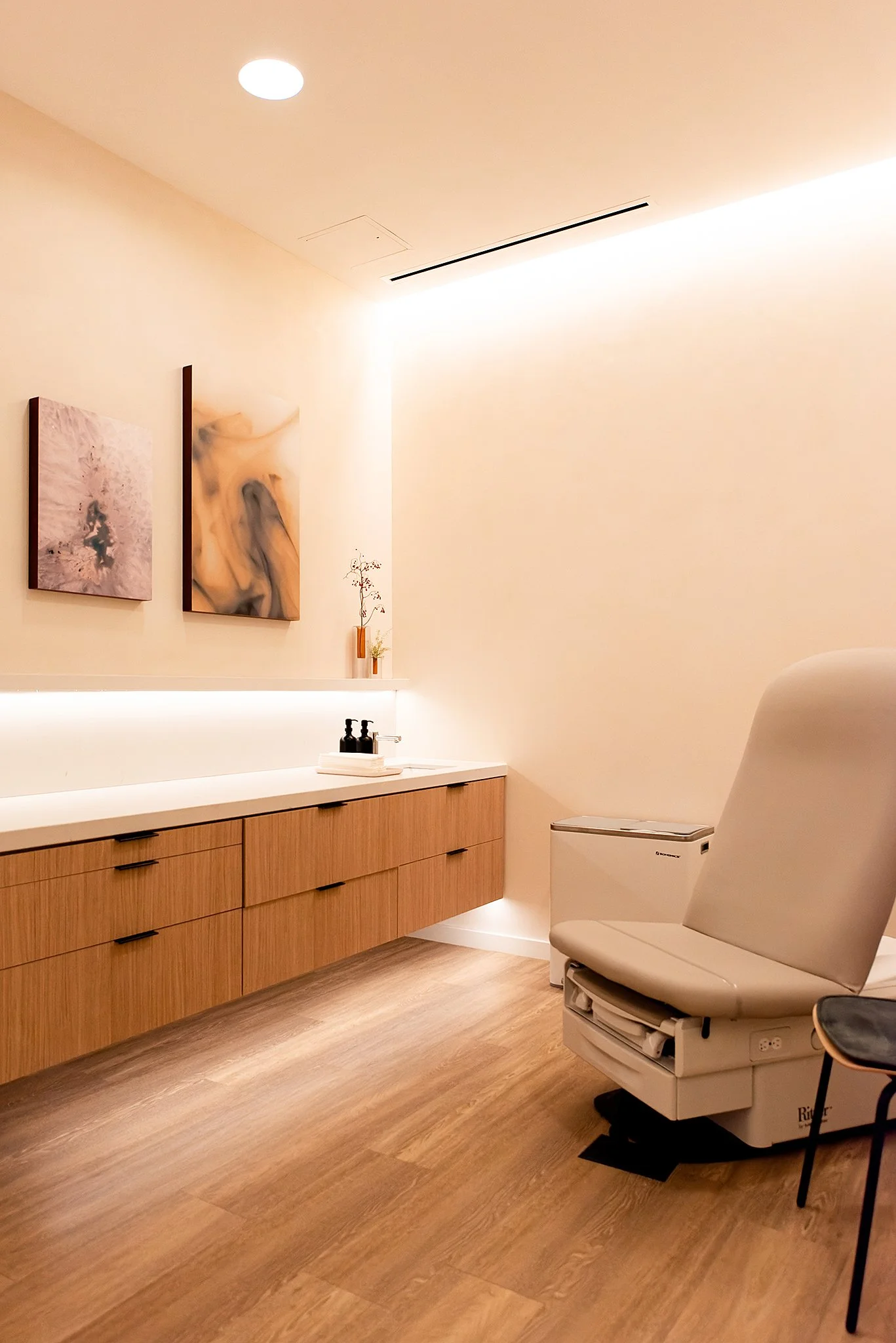 A modern, minimalist room with wooden flooring, a wooden cabinet with black handles, abstract wall art, a chair, and a white medical examination table.