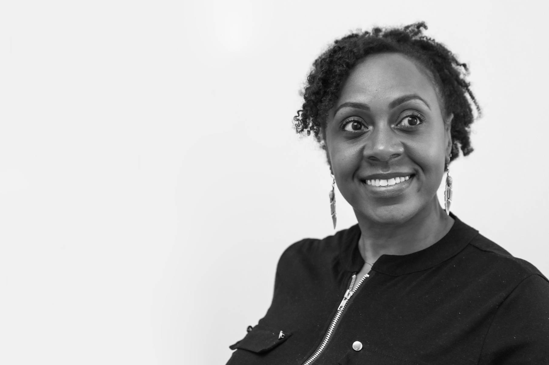 Black and white photo of a smiling woman with curly hair, wearing earrings and a zip-up shirt, looking to her right.