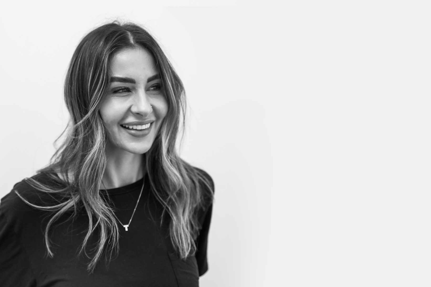 Black and white photo of a smiling woman with long wavy hair, wearing a black top and a necklace with a T-shaped pendant, against a plain light background.